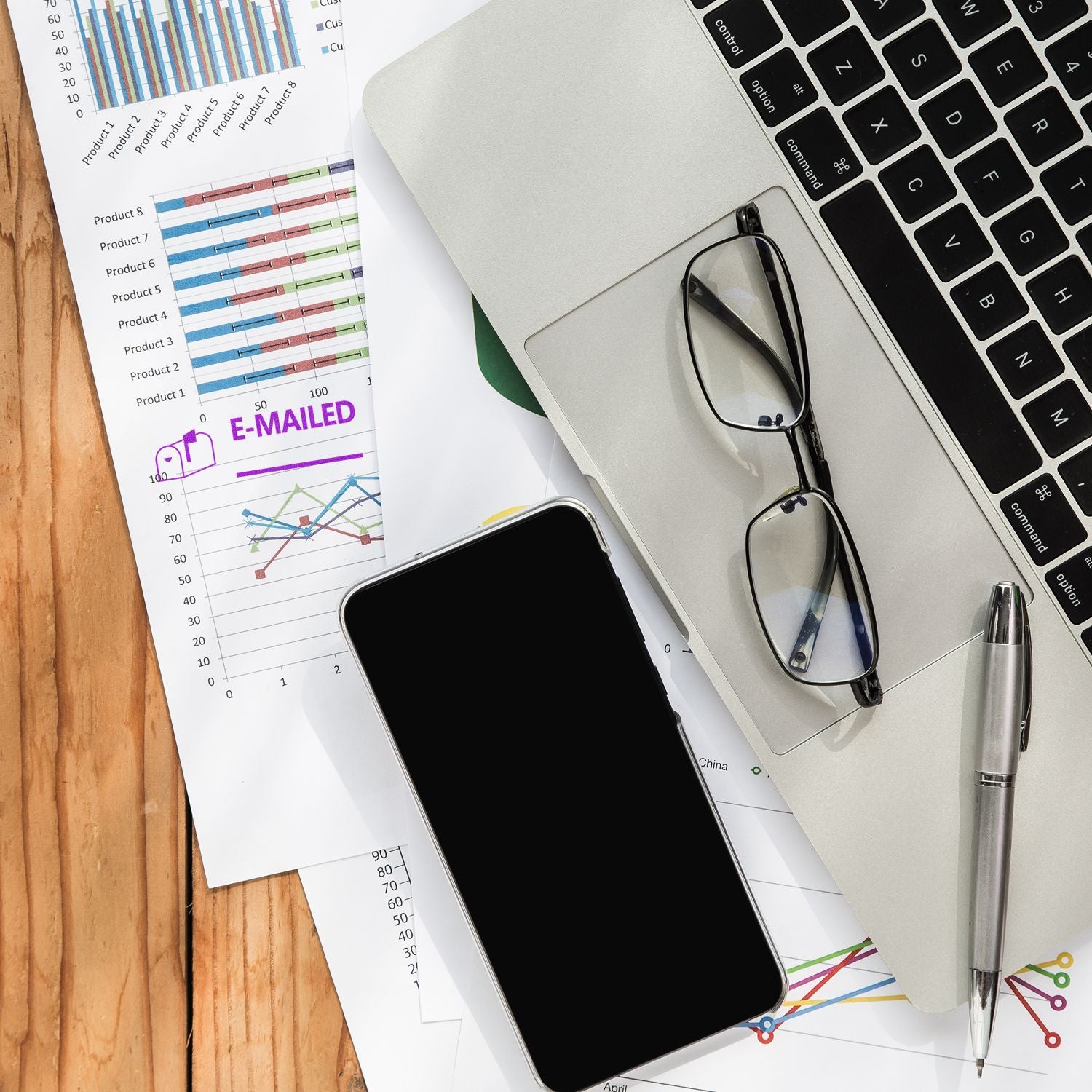Business analytics report with colorful charts labeled E-MAILED, on desk beside laptop keyboard, smartphone, and glassesdata tracking.
