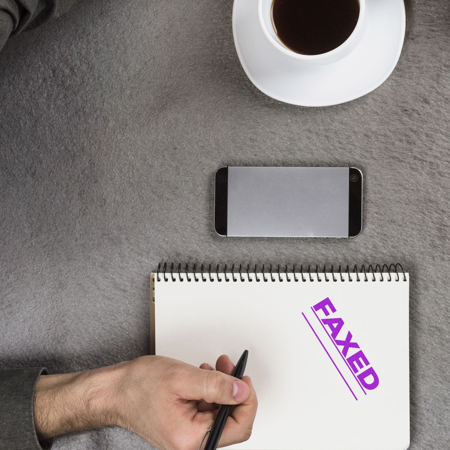 Business desk with coffee, smartphone, and notebook stamped FAXED as a hand writes with a pen.
