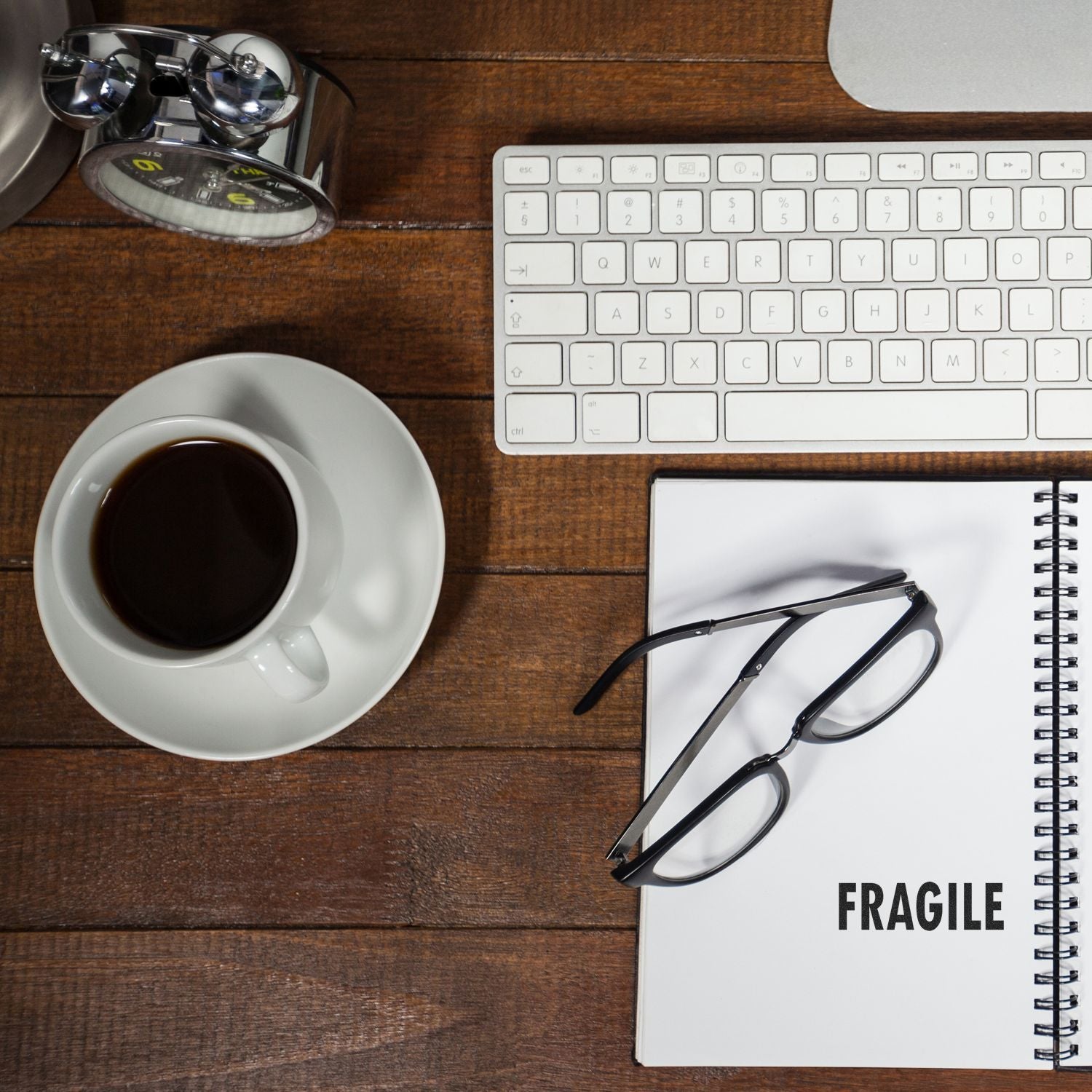 Top view of office desk with coffee cup, keyboard, eyeglasses, alarm clock, and notebook labeled fragile