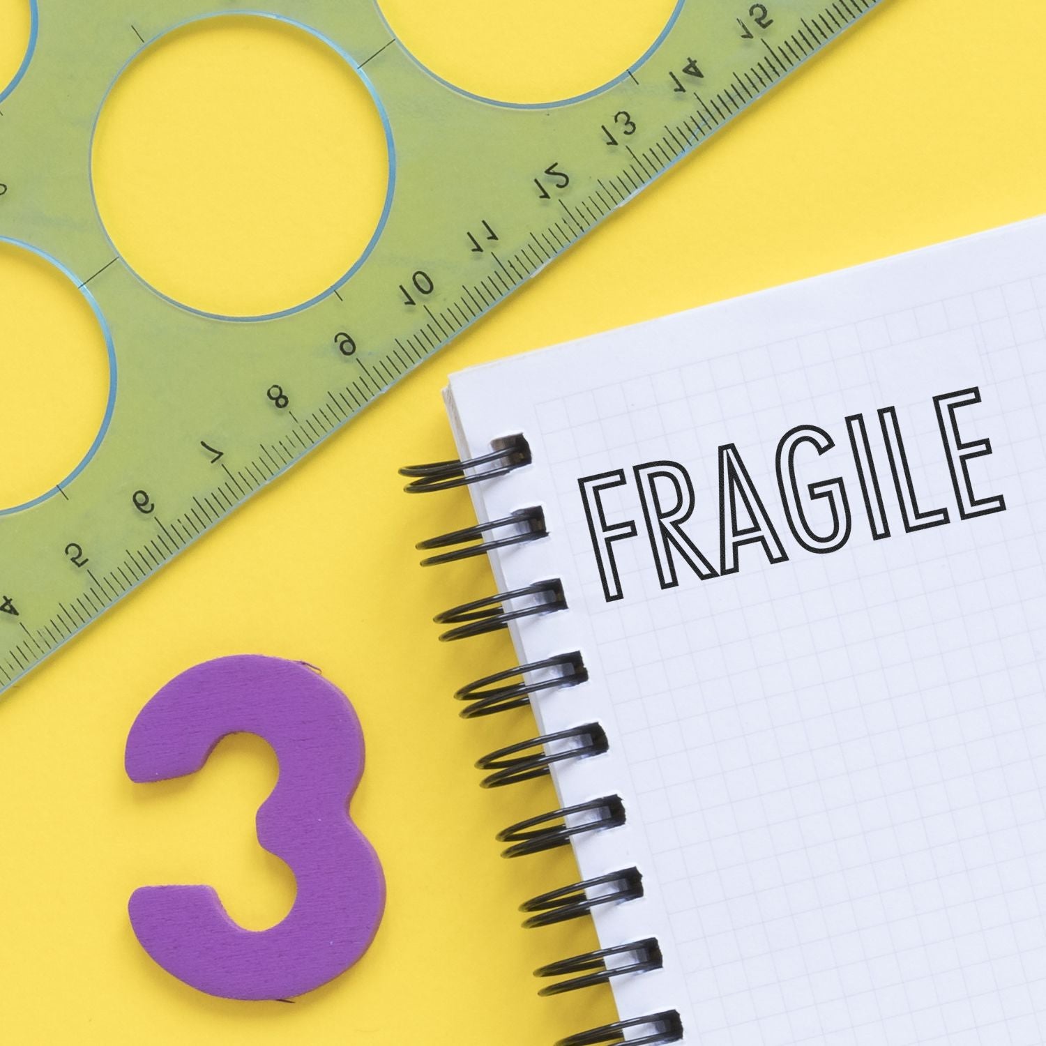Fragile text in a spiral notebook with ruler, stencil, and number 3 on a bright yellow background.