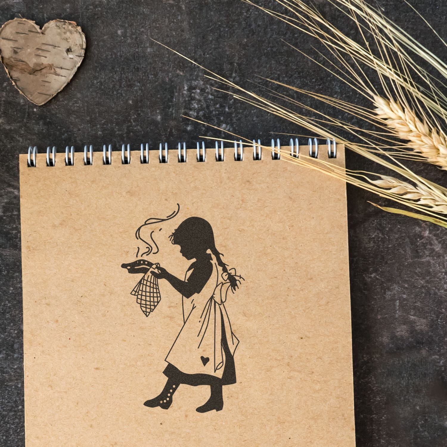 Brown spiral notebook with a girl silhouette holding a steaming cup, styled on a rustic flat lay with wheat stalks and a birch heart decoration.
