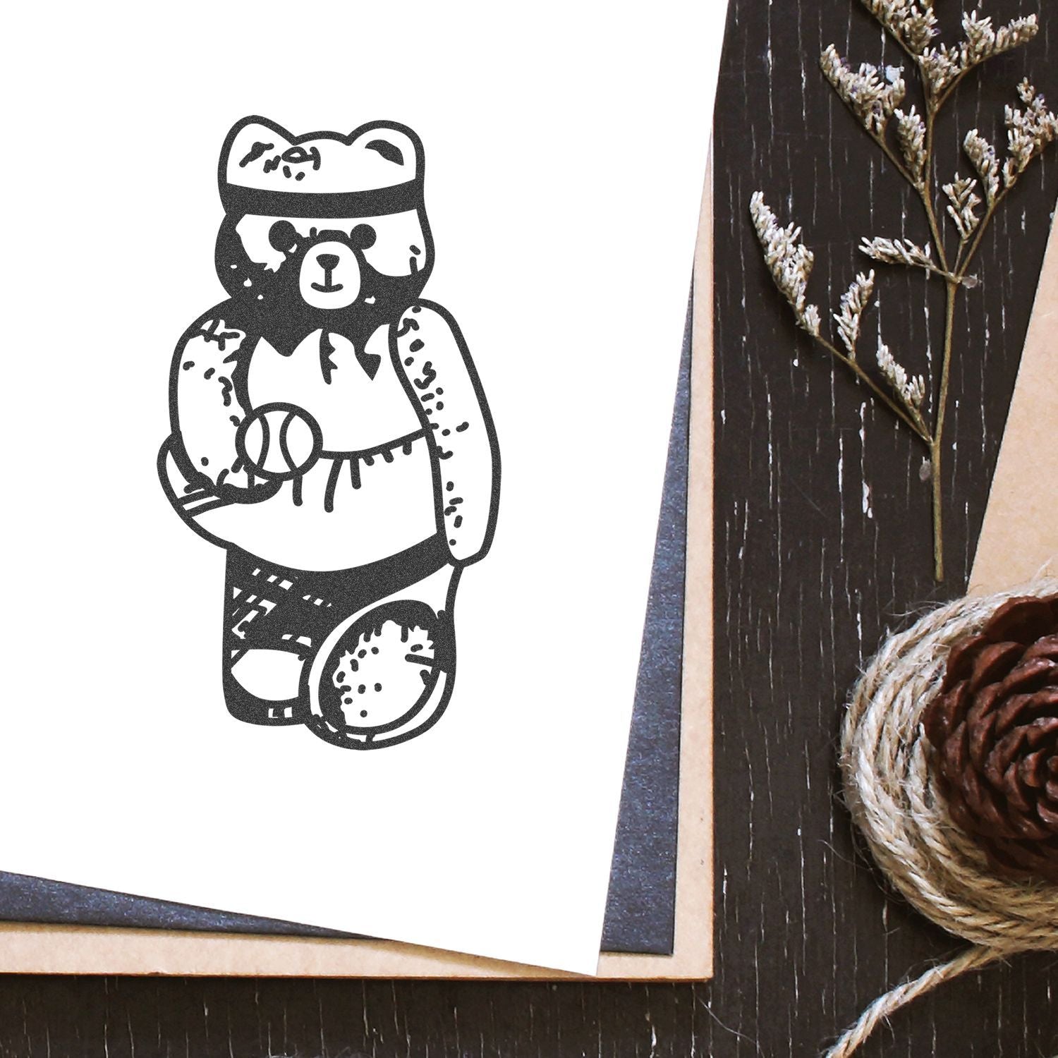 Cute black bear illustration on paper with rustic pinecone, twine, and dried floral decor on a wooden background.