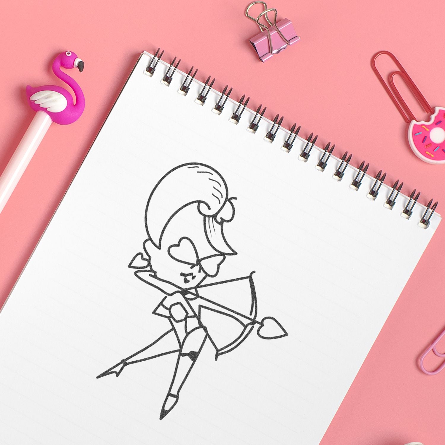 Pink stationery flat lay with spiral notebook, cute Cupid doodle, flamingo pen, donut paper clips, and binder clip on pastel pink background.