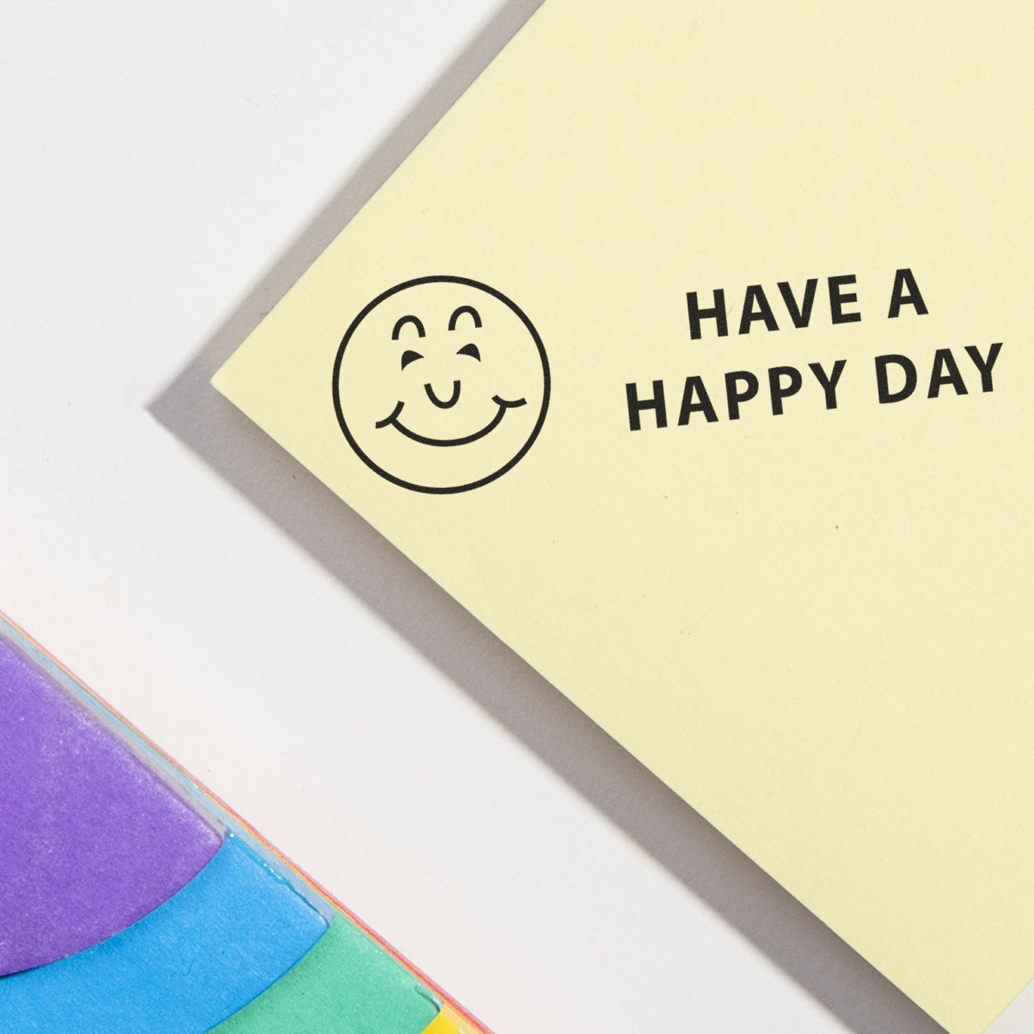 Smiling sticky note with Have a Happy Day text and colorful rainbow paper on a white background.