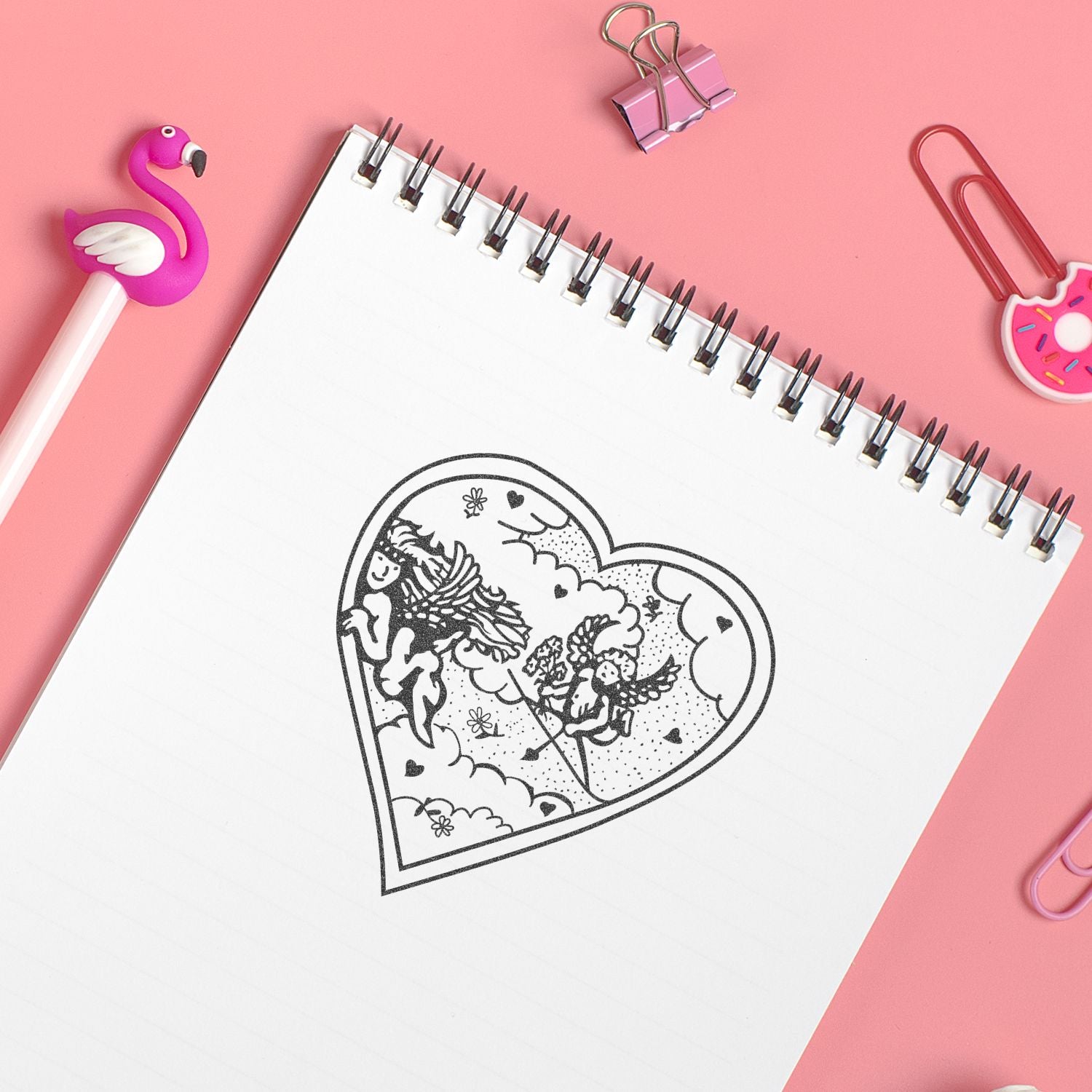 Pink desk flat lay with a spiral notebook showing a heart-shaped angel doodle, surrounded by cute stationery and paper clips.