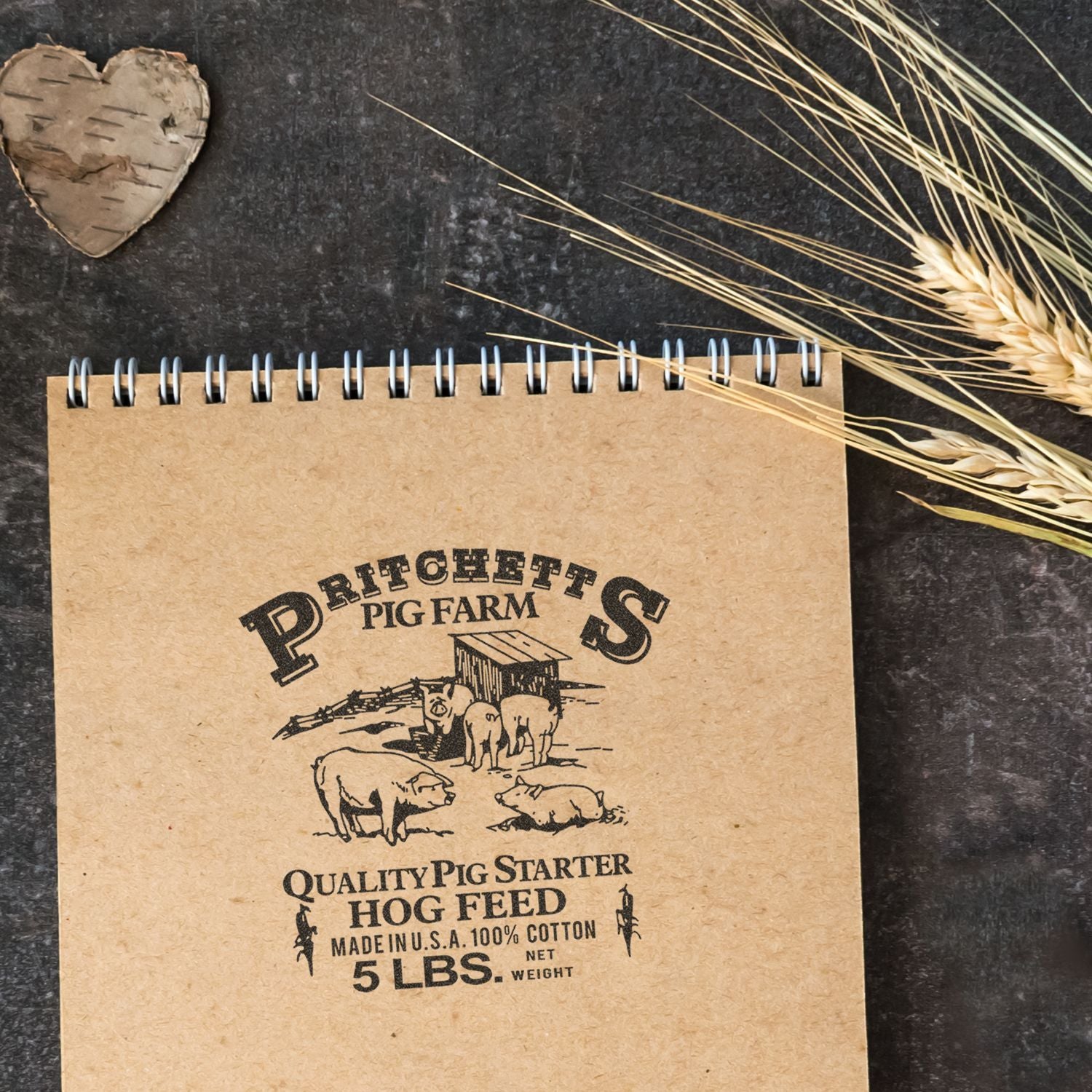 Rustic spiral notebook with vintage Pritchetts Pig Farm hog feed graphic, styled with dried wheat on a dark background.
