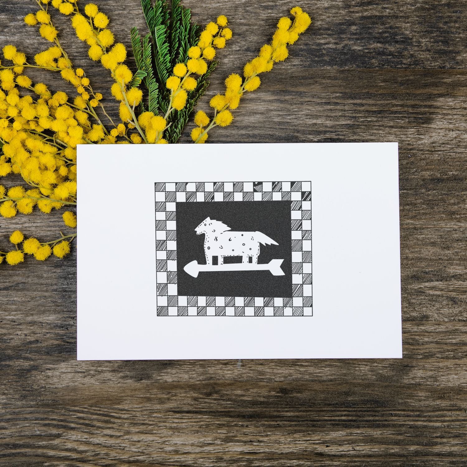 Yellow mimosa flowers on rustic wood beside a white card with a black-and-white horse illustration.