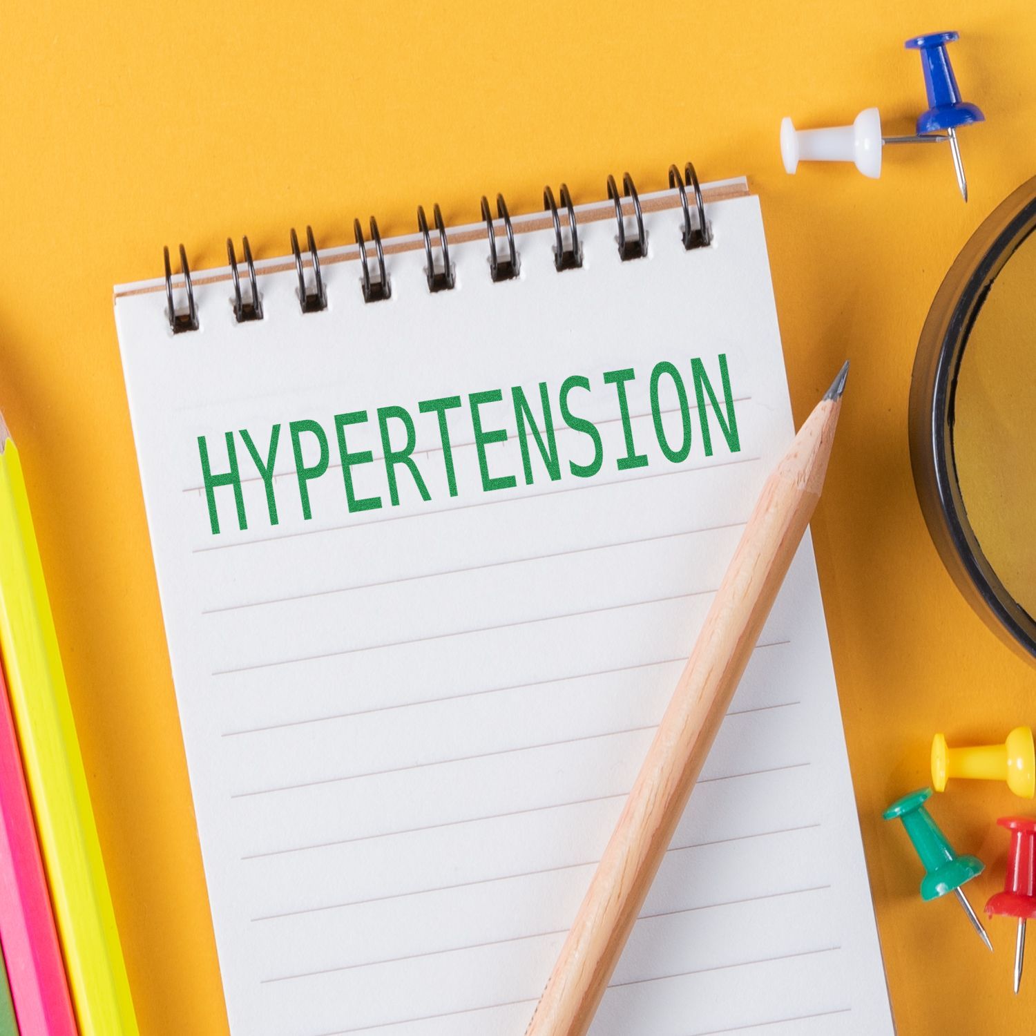Notepad with hypertension text, pencil, and colorful push pins on a yellow background, health concept.