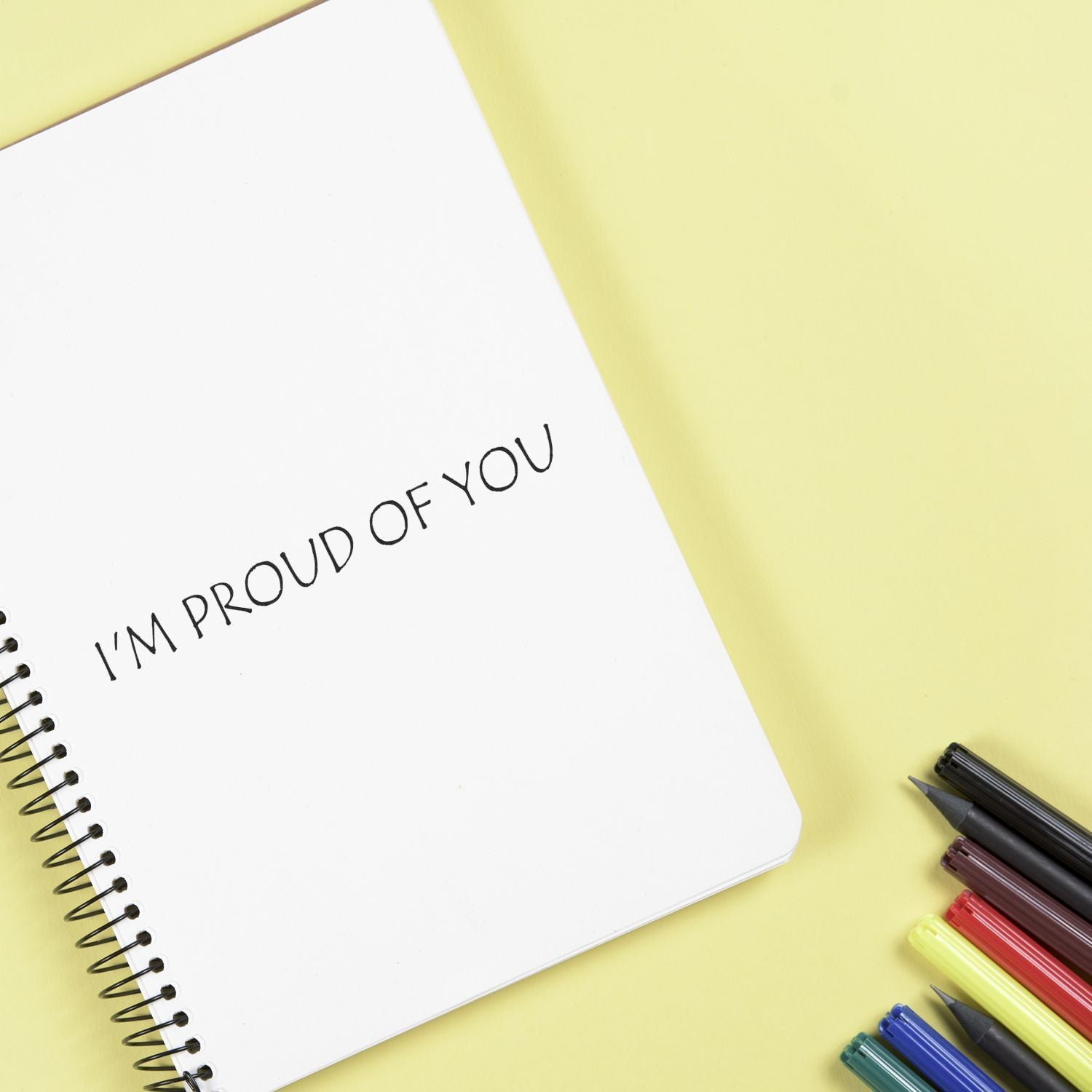 Motivational note in open notebook reading Im proud of you with colorful pens on a yellow desk