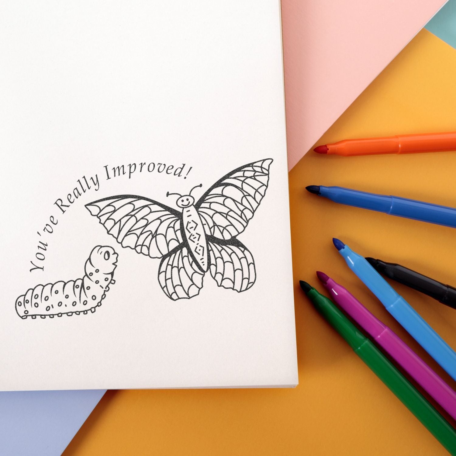 Open coloring book page with butterfly and caterpillar doodle reading Youve Really Improved! surrounded by colorful markers on a pastel background.