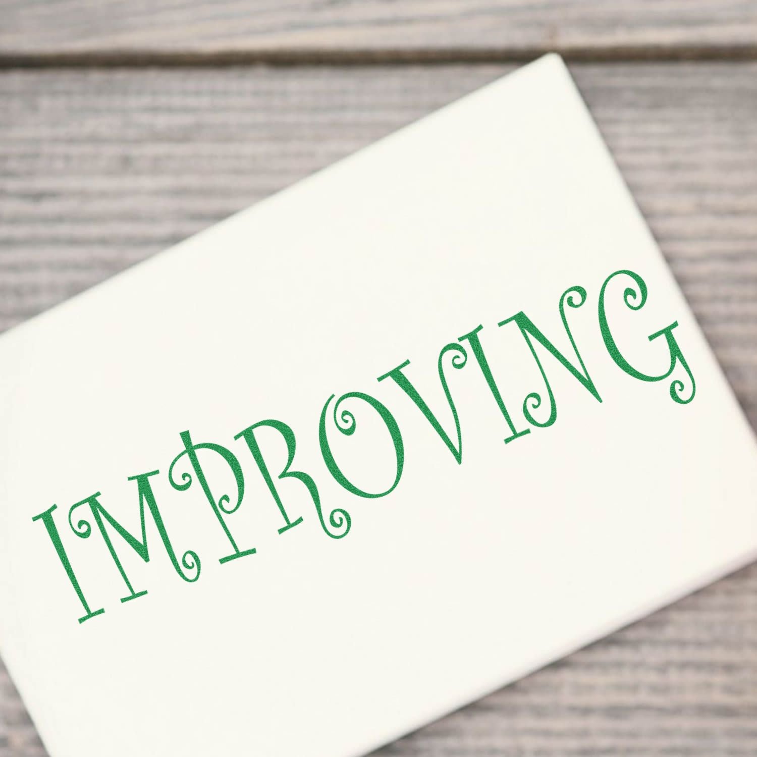 White greeting card with green IMPROVING text on a rustic wood background