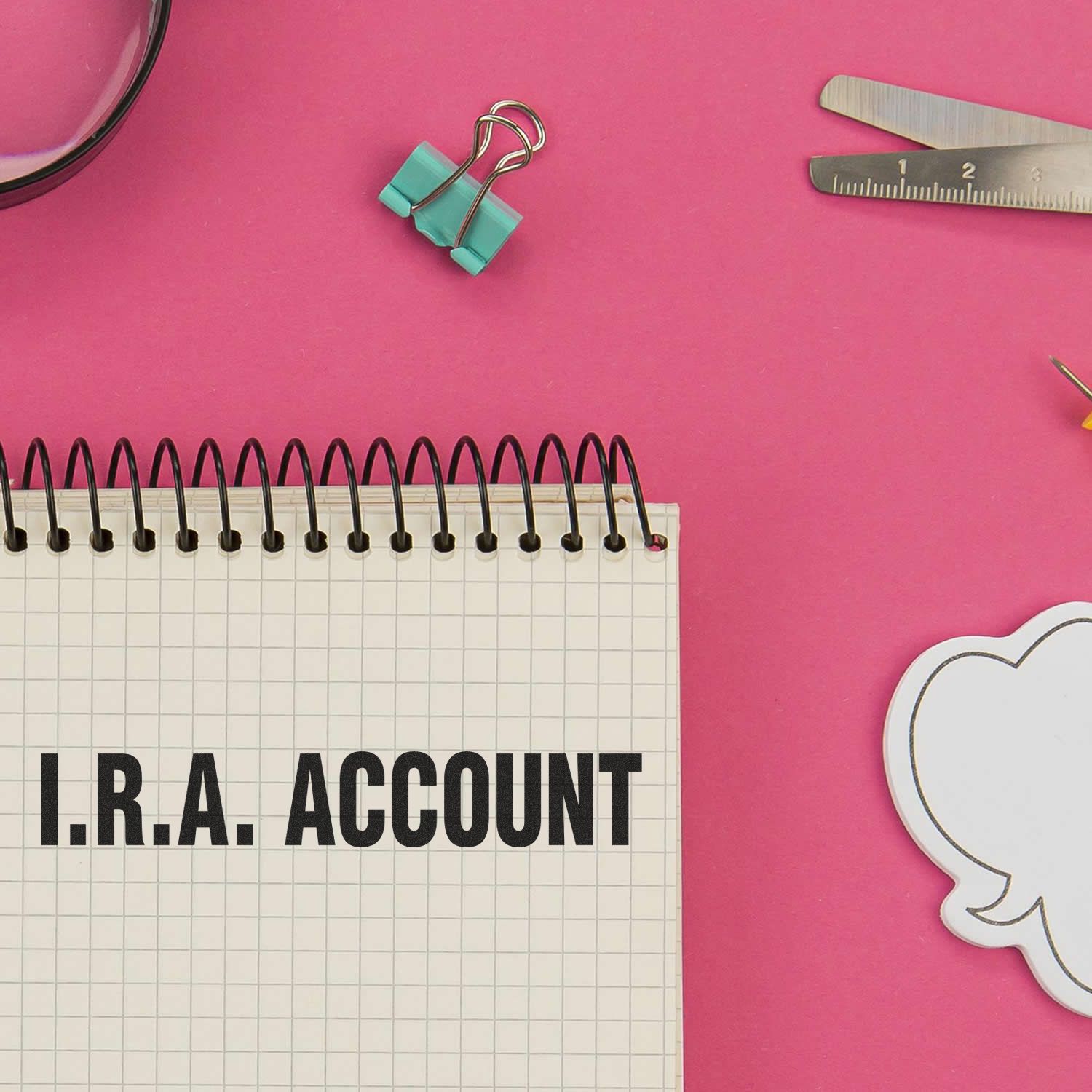 Top view of I.R.A. ACCOUNT notebook with binder clips and ruler on pink background, showing IRA retirement accounting