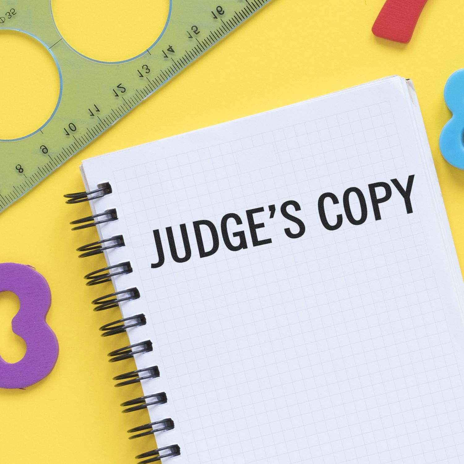 Notebook page labeled JUDGES COPY on a colorful school supply table with ruler, crayons, and learning tools for class worksheet use.