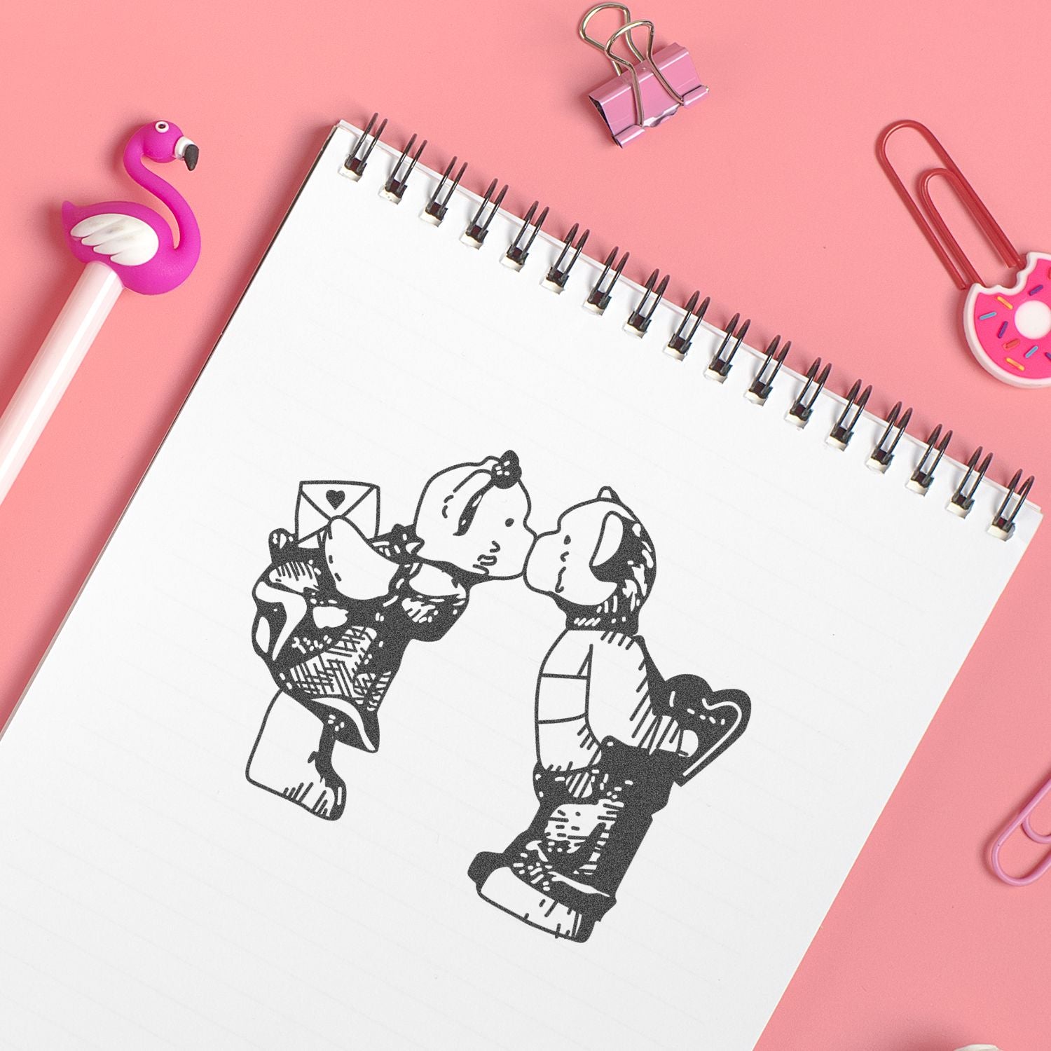 Cute spiral notebook with black doodle illustration, surrounded by pink stationery accessories on a pastel pink background.