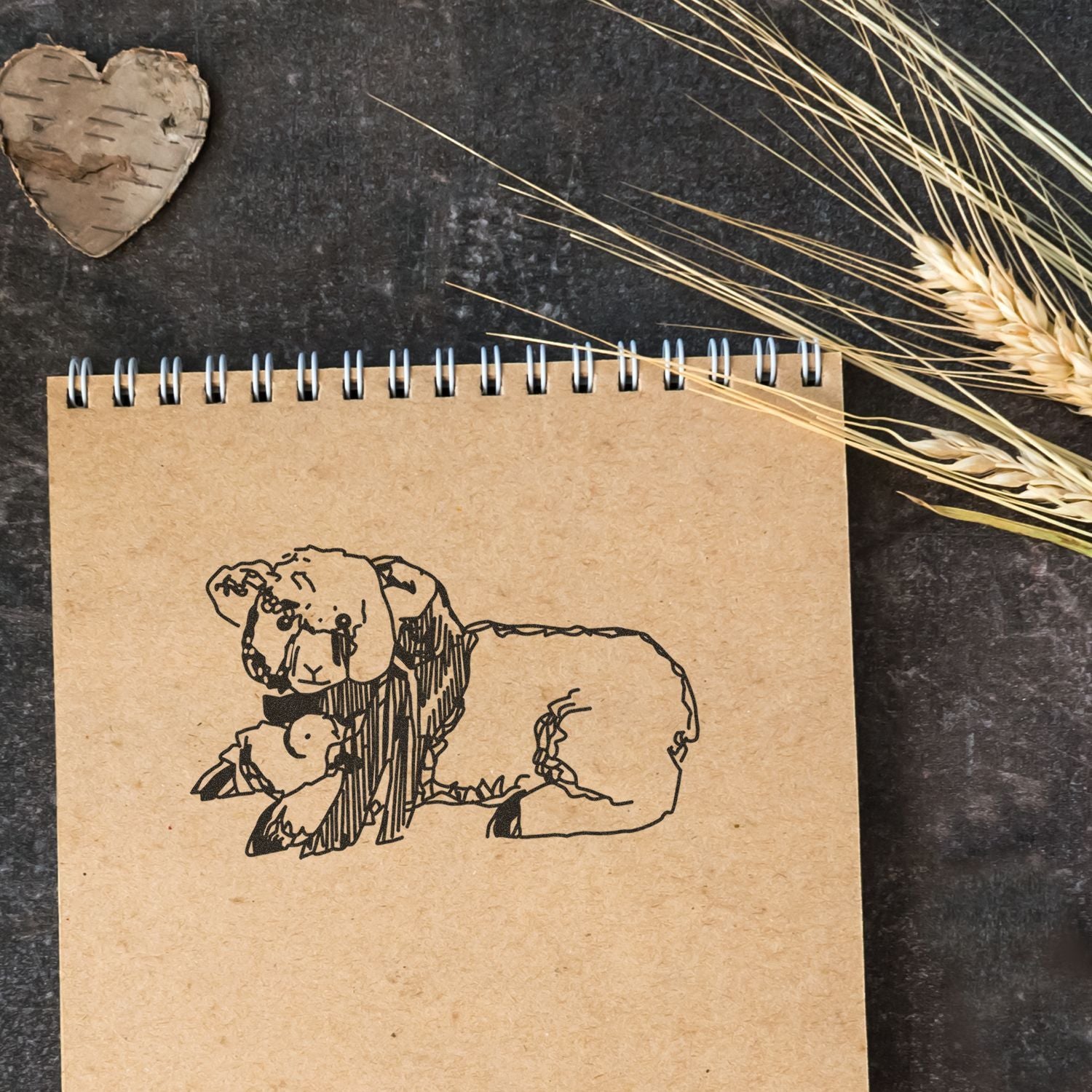 Rustic spiral notebook with a hand-drawn sheep illustration, wheat stalks, and a wooden heart on a dark textured background.