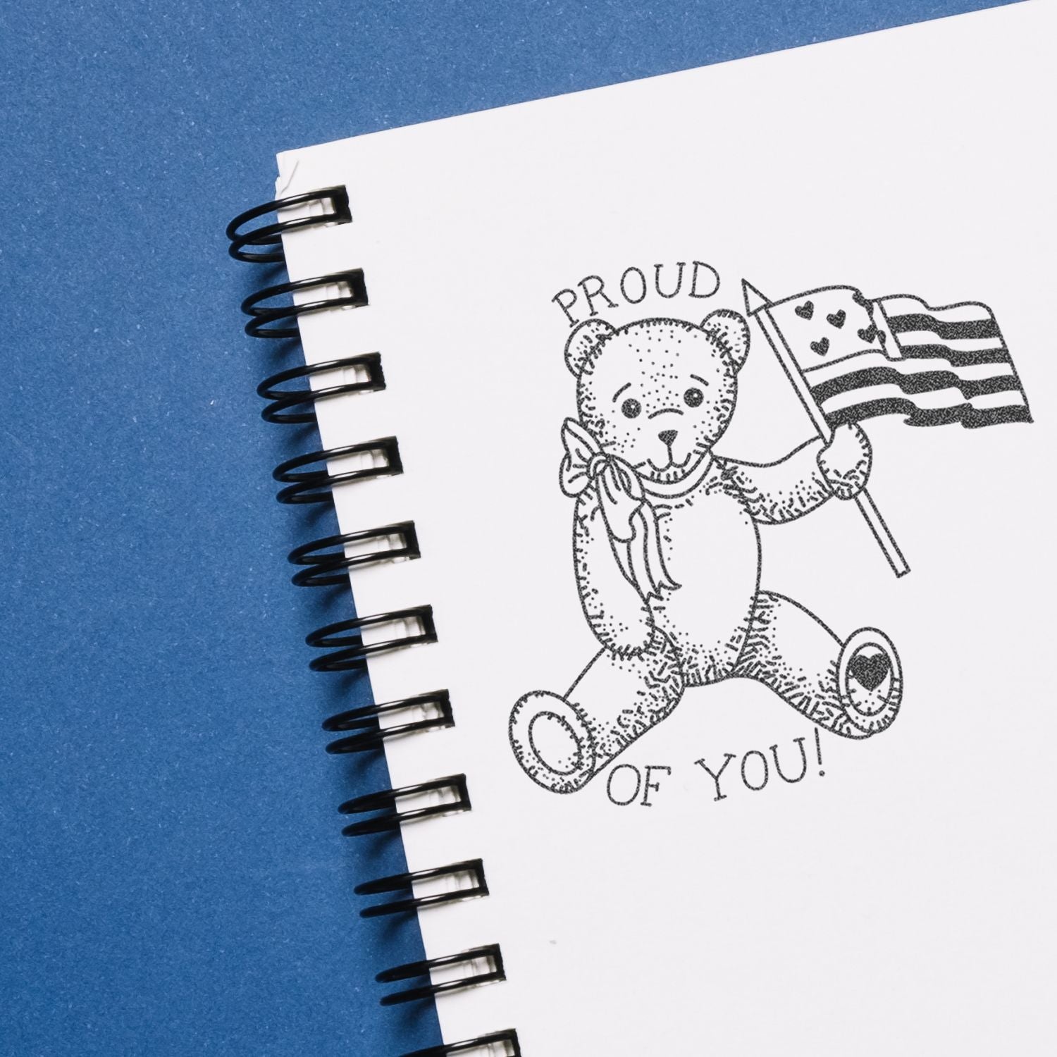Spiral notebook page with teddy bear holding a pride flag and Proud of You! message on a blue background.