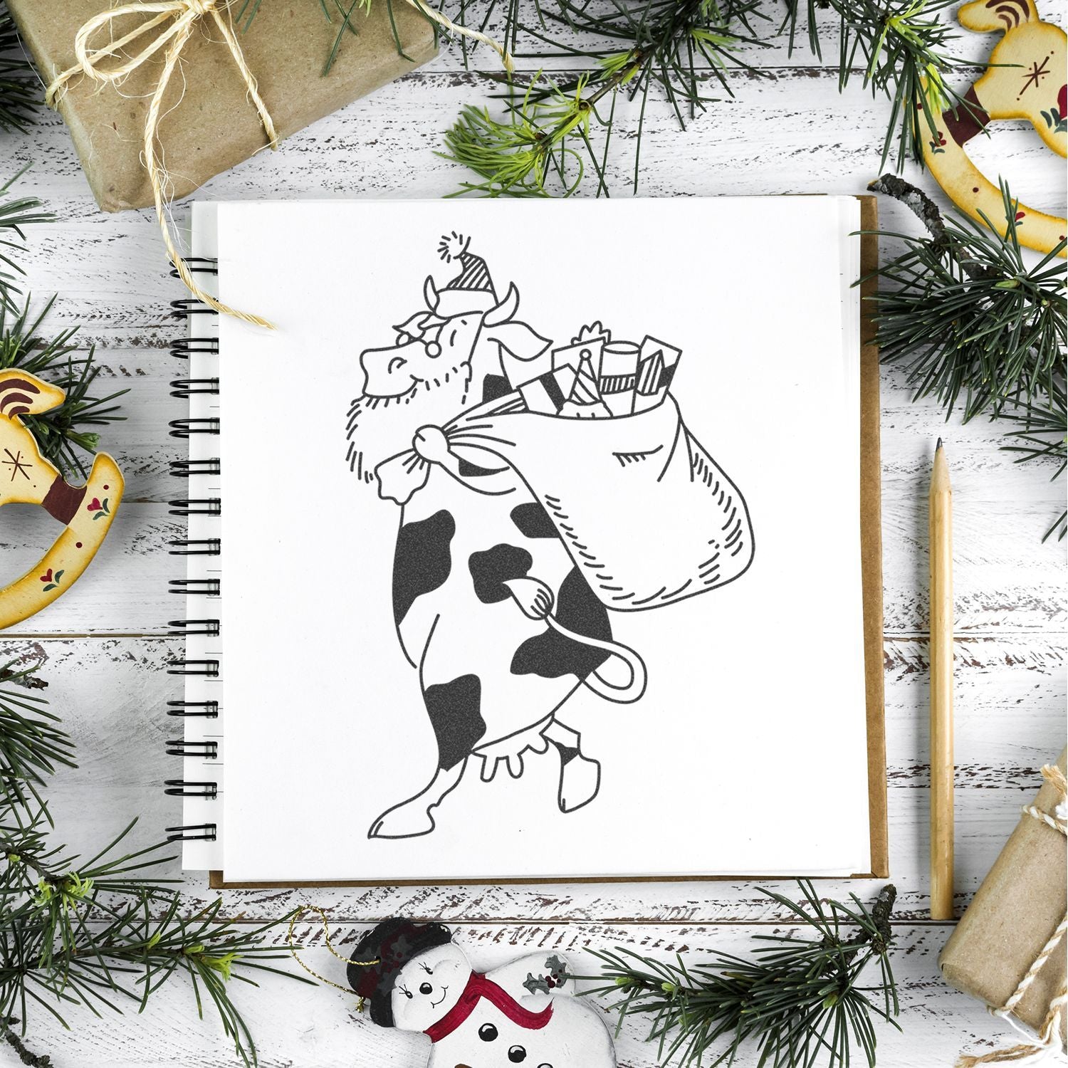 Christmas cow illustration in a sketchbook surrounded by festive pine branches, wrapped gifts, and holiday decorations on a rustic white wooden table