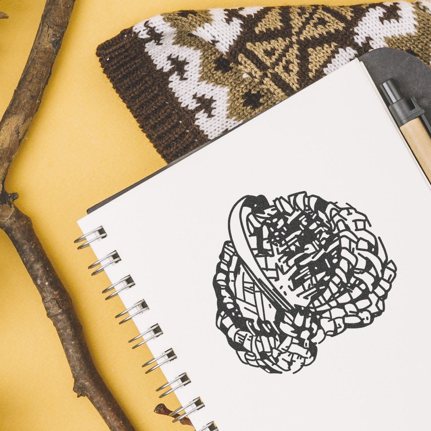 Spiral notebook with abstract black line art, pen, knitted fabric, and natural wood sticks on a yellow background.
