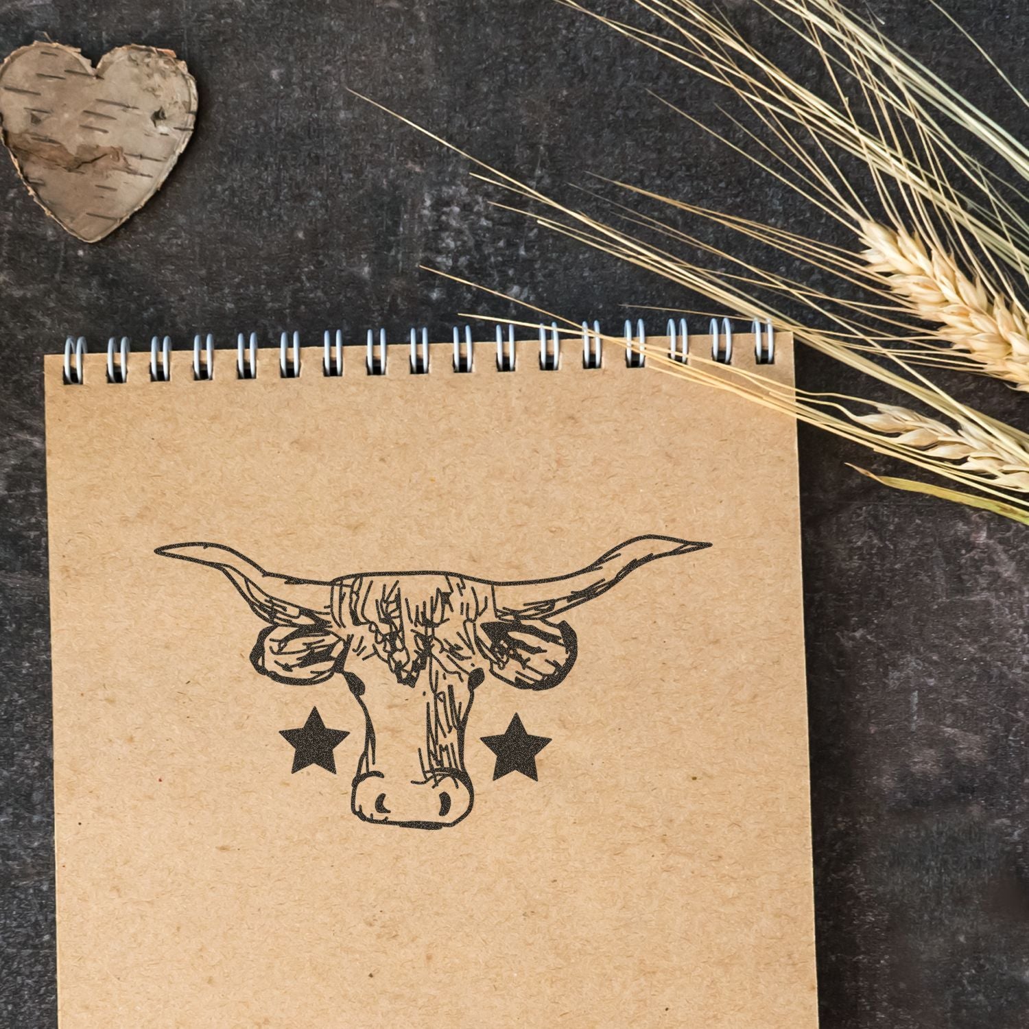 Rustic spiral notebook with longhorn skull graphic and stars on kraft paper, styled with dried wheat and birch heart on a dark background.