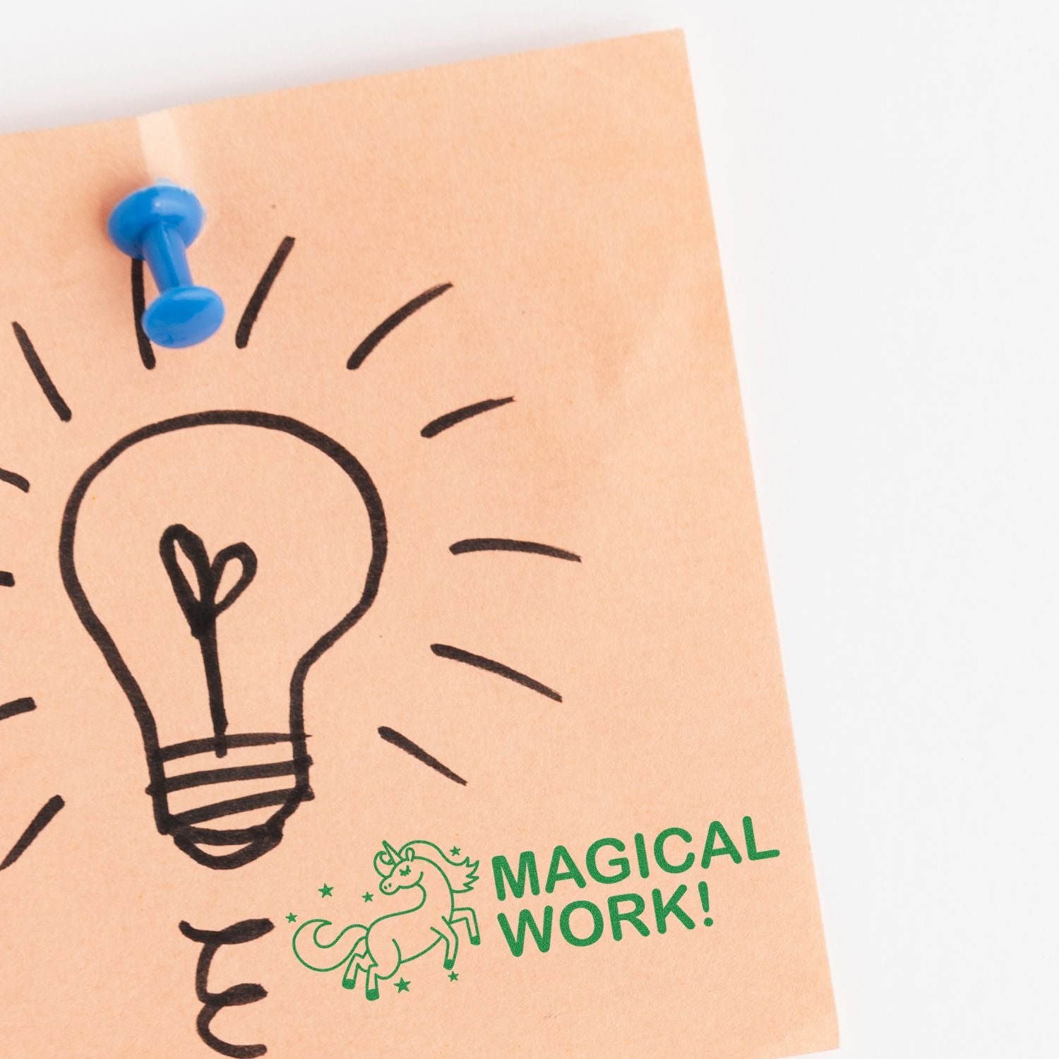 Blue pushpin holds a motivational note reading Magical Work! with a light bulb doodle on kraft paper.