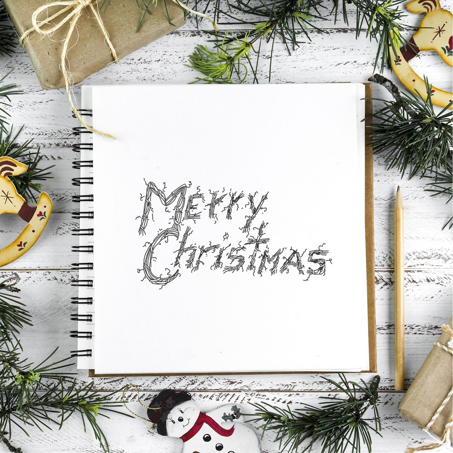 Open sketchbook with hand-drawn Merry Christmas lettering, festive pine branches, wrapped gifts, and holiday decor flat lay.
