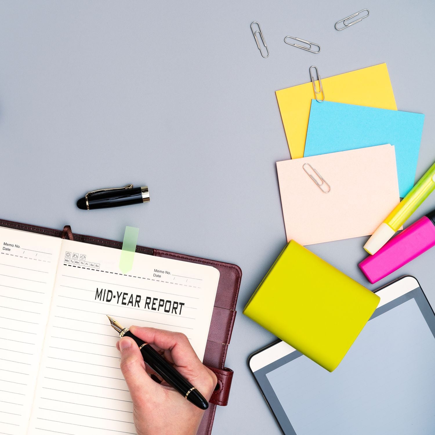 Hand writing a mid-year report in a notebook on an office desk with colorful sticky notes, pens, and planning supplies for business review.