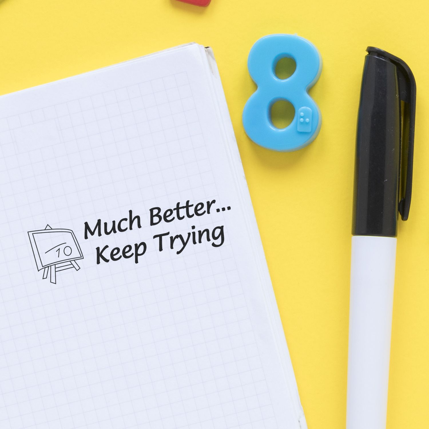 Motivational quote on notebook: Much Better, Keep Trying, with pen and blue number 8 eraser on yellow office desk background.