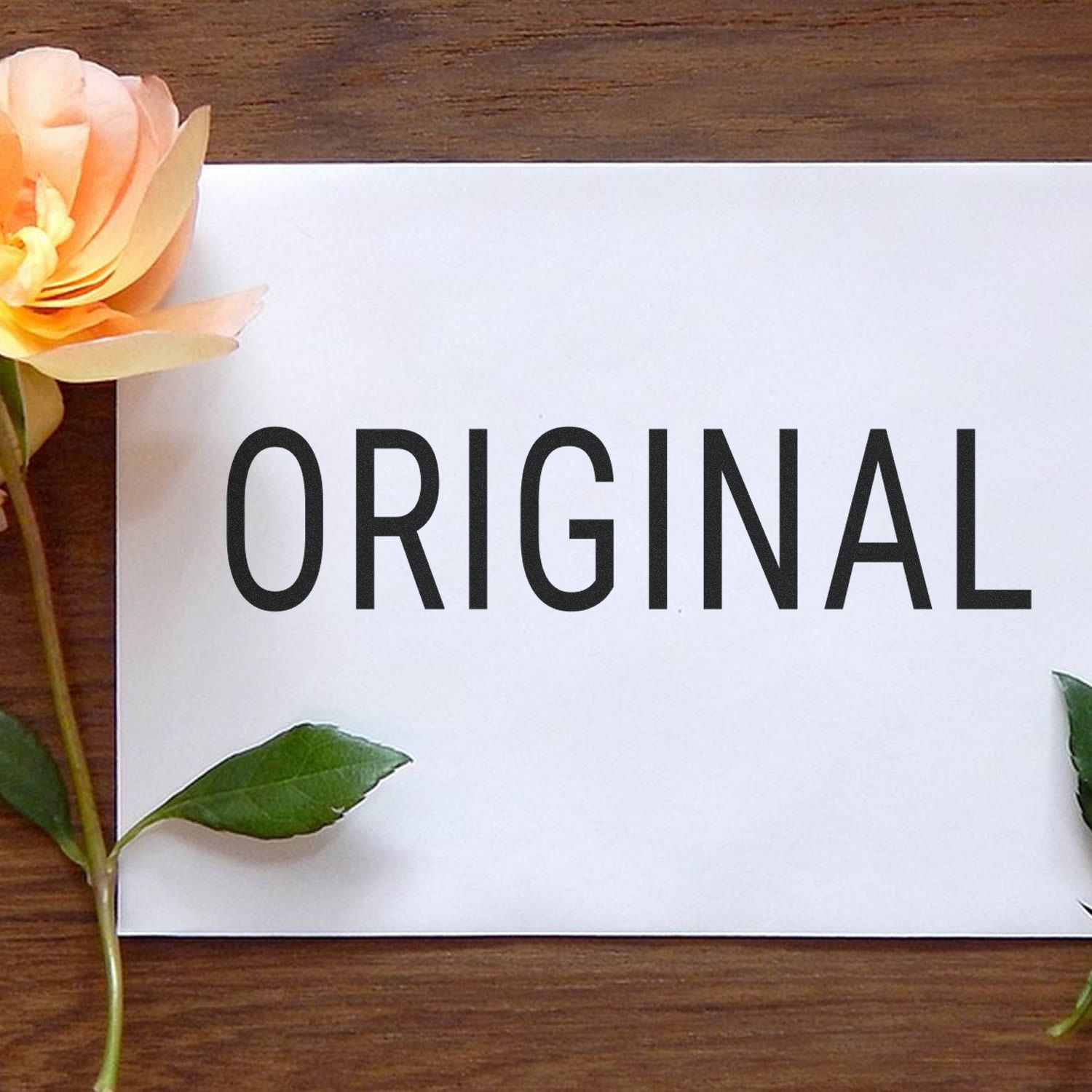 White paper with the word ORIGINAL on a wooden table, decorated with a peach flower and green leaves.