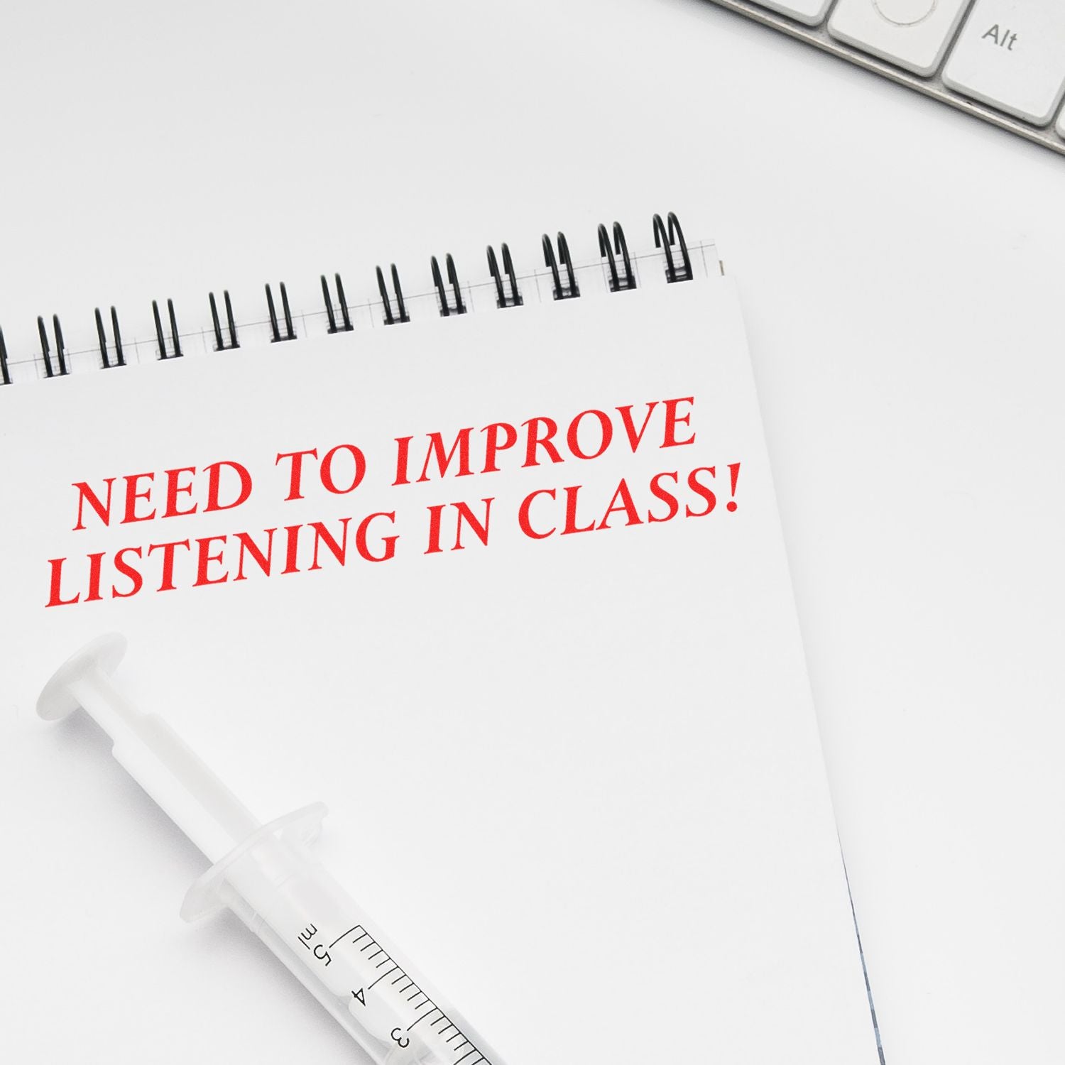 Notebook with red text Need to improve listening in class! beside a syringe on a white desk with a keyboard.