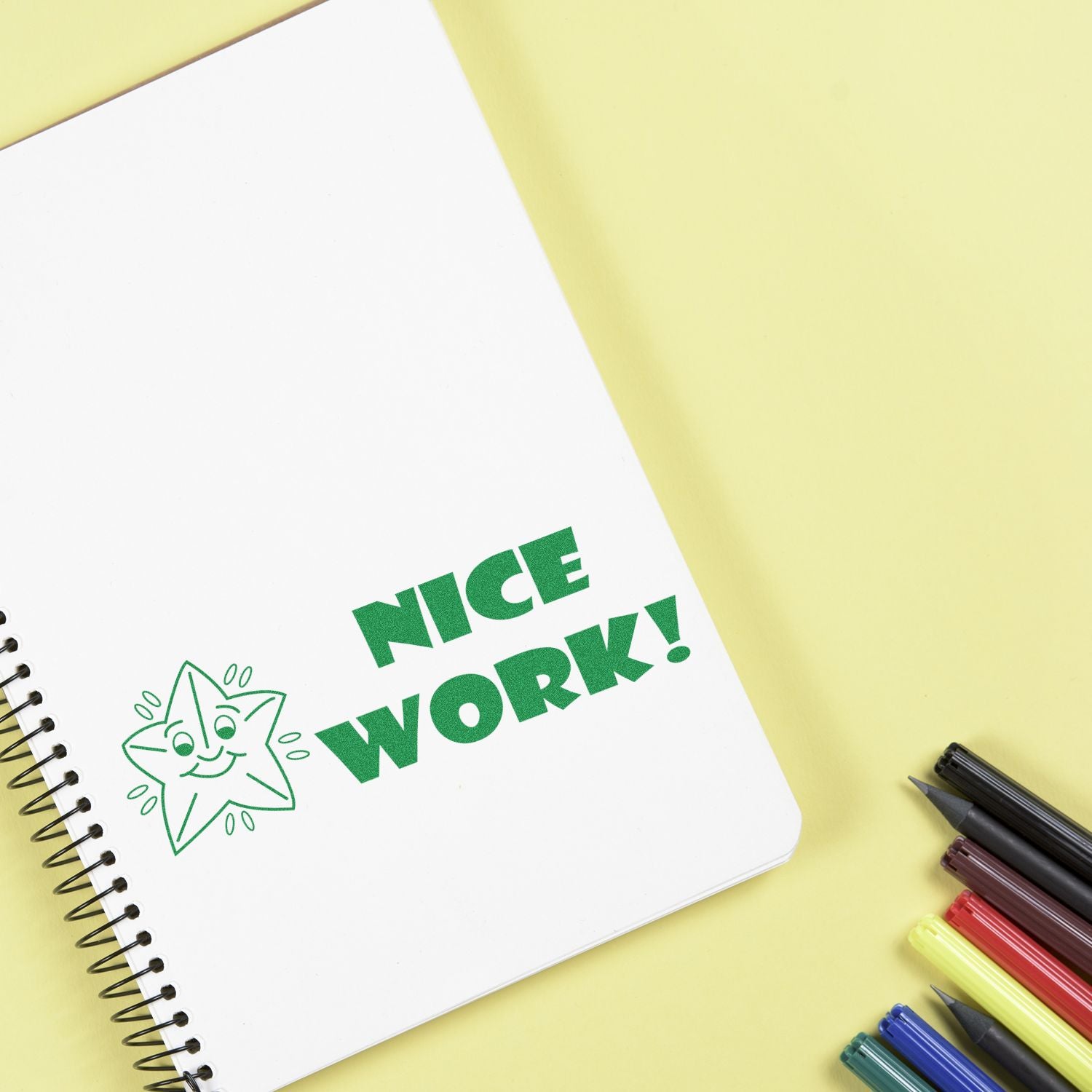 White spiral notebook with Nice Work! text and smiling star illustration on a yellow background, surrounded by colorful pens.