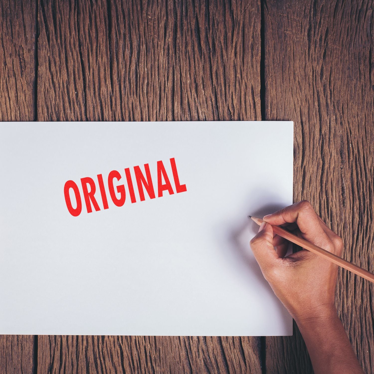 Hand writing ORIGINAL on paper with pencil, conveying authenticity, creativity, and original branding message on wood background.
