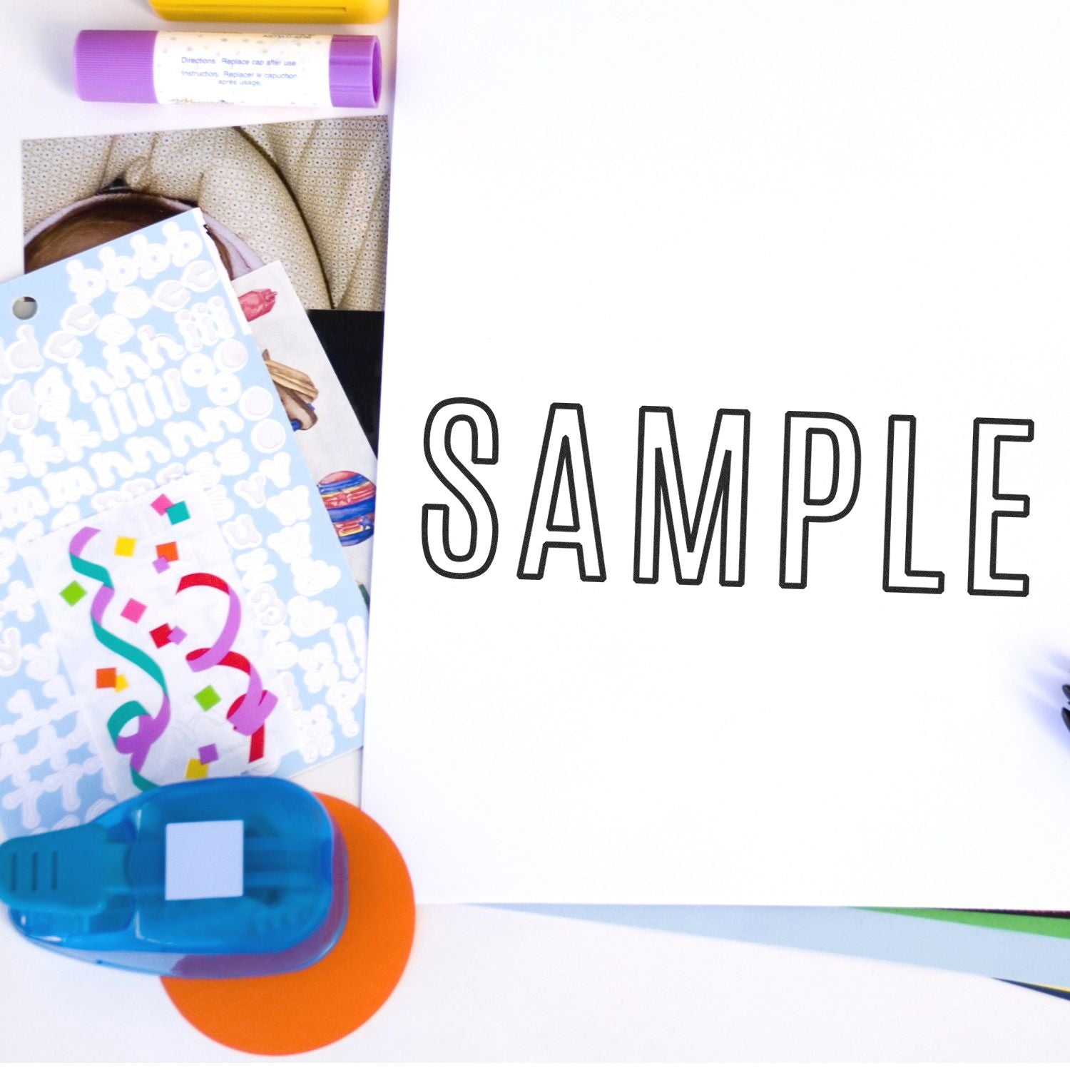 Scrapbooking supplies and colorful paper crafts with a SAMPLE page and adhesive tape dispenser on a white background.