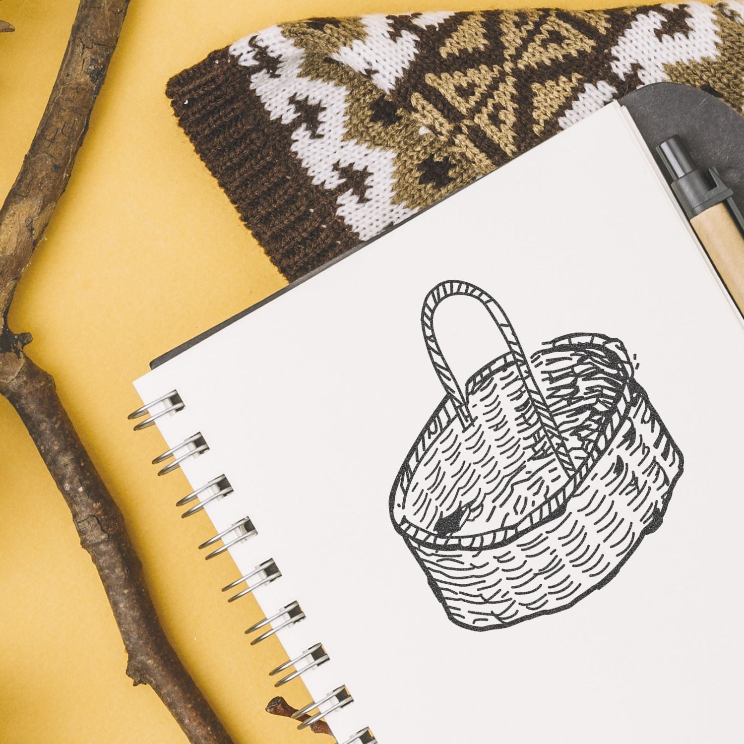 Sketchbook with hand-drawn woven basket illustration, pen, rustic branches, and knitted textile on a yellow background.