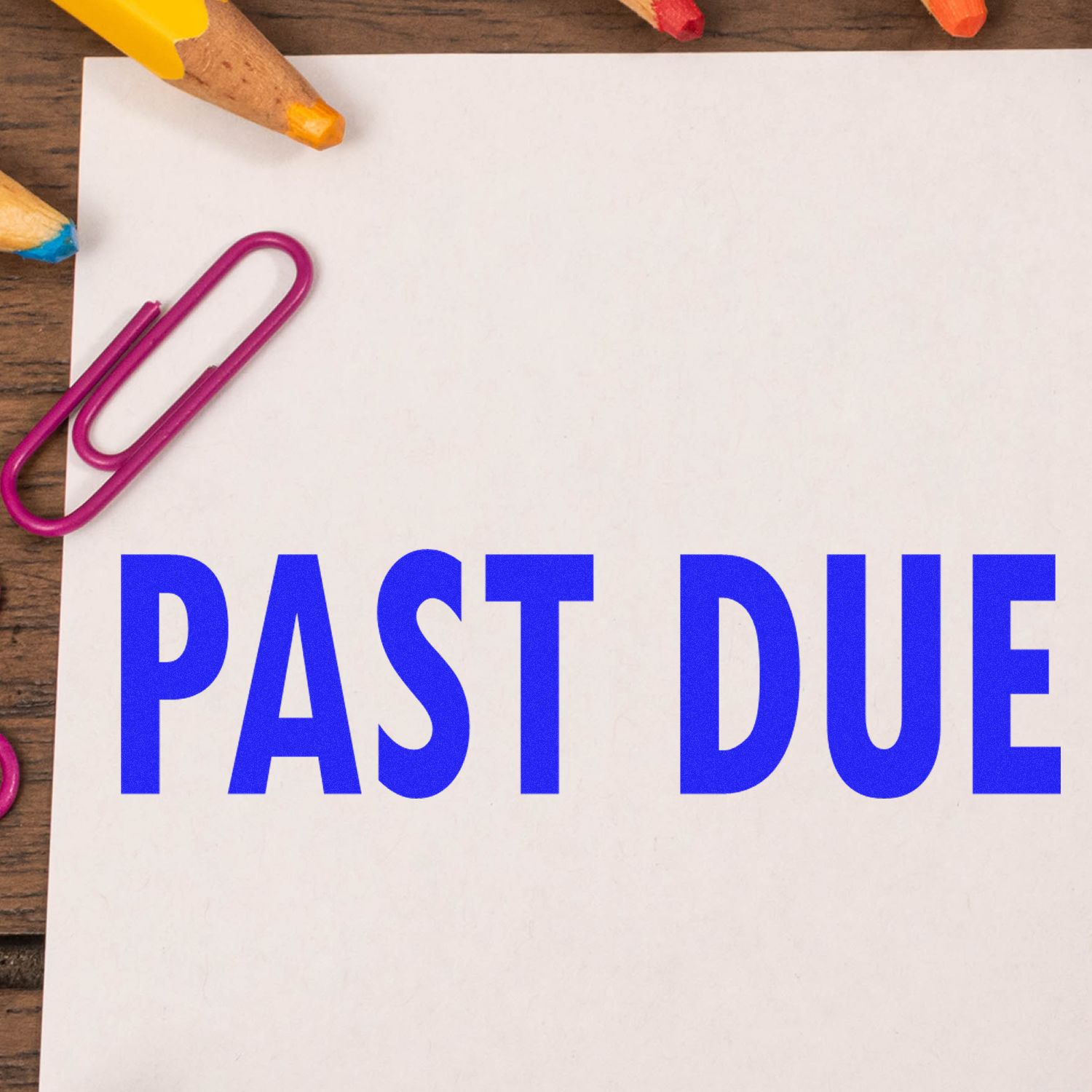 Past due reminder note on white paper with colored pencils and paper clip on wooden desk.