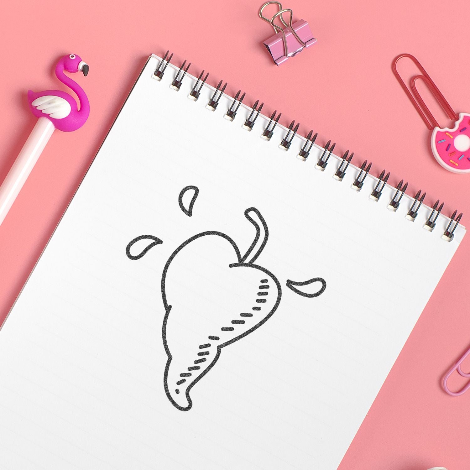 Pink stationery flat lay with a spiral notebook featuring a hand-drawn fruit sketch, flamingo pen, and colorful paper clips.