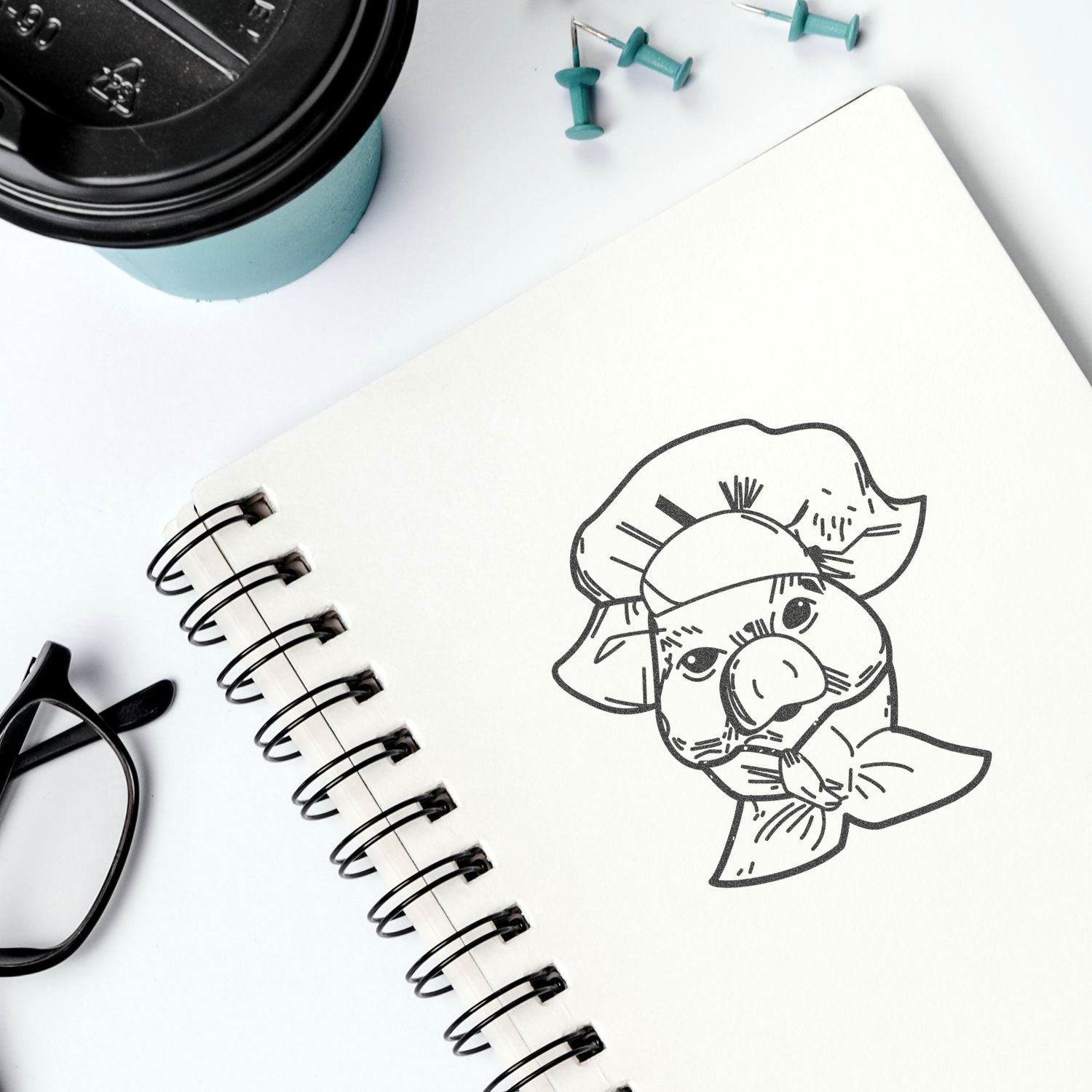 Sketchbook with pig illustration on a white desk beside a coffee cup, eyeglasses, and push pins.