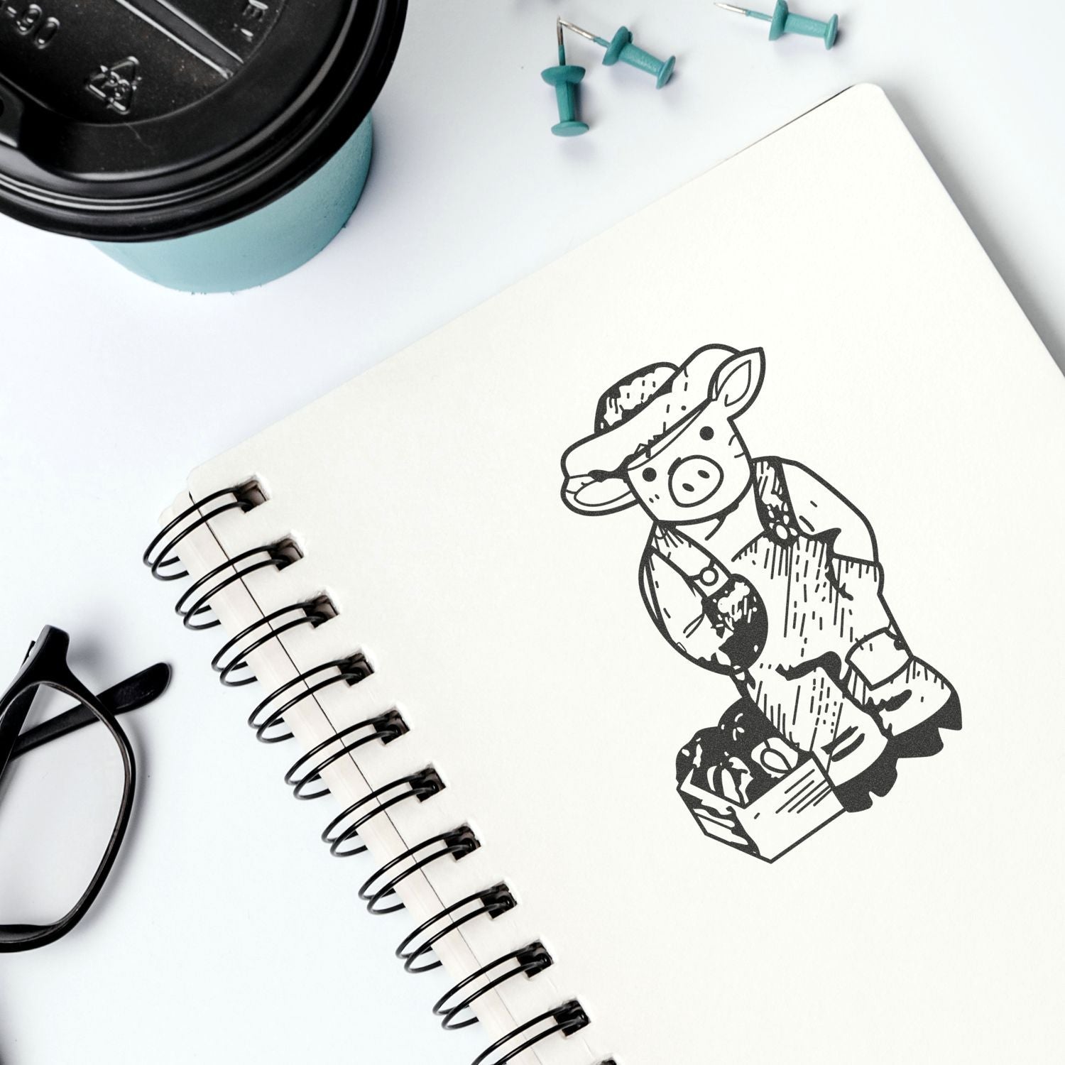 Spiral notebook with pig illustration, coffee cup, glasses, and push pins on a white desk, top view.