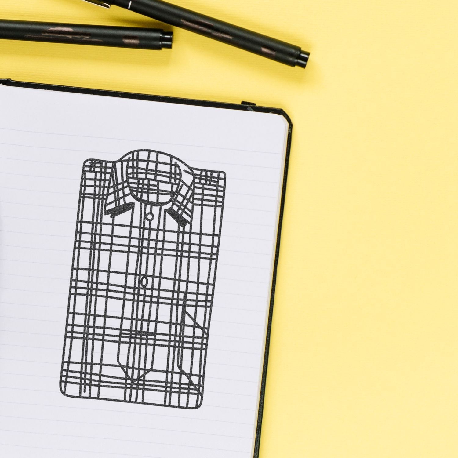 Fashion design sketch of a plaid button-up shirt in a notebook on a yellow background with black pens.