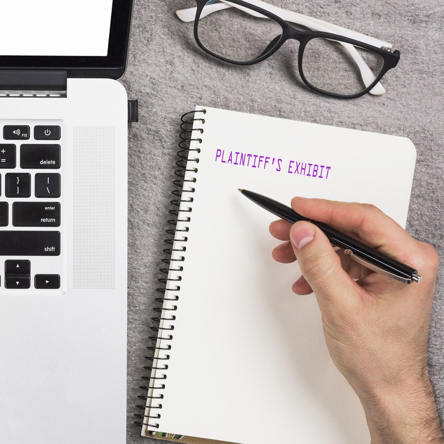 Hand writing Plaintiffs Exhibit in a notebook beside a laptop and eyeglasses on a desk.