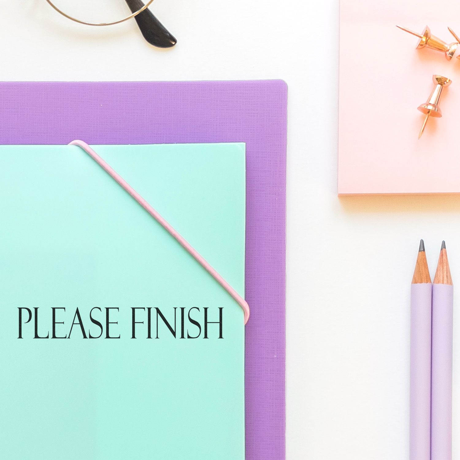 Pastel stationery flat lay with Please Finish notebook, pencils, push pins, and eyeglasses on a white desk background