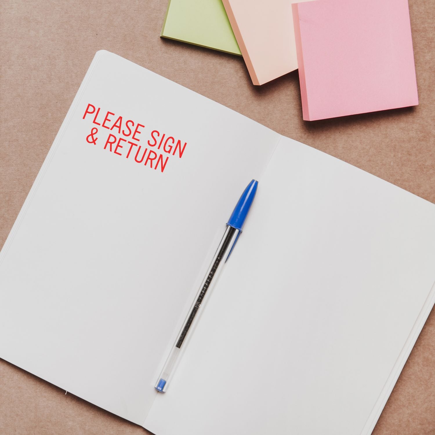 Open notebook with Please Sign & Return text, blue pen, and pastel sticky notes on a brown desk background.