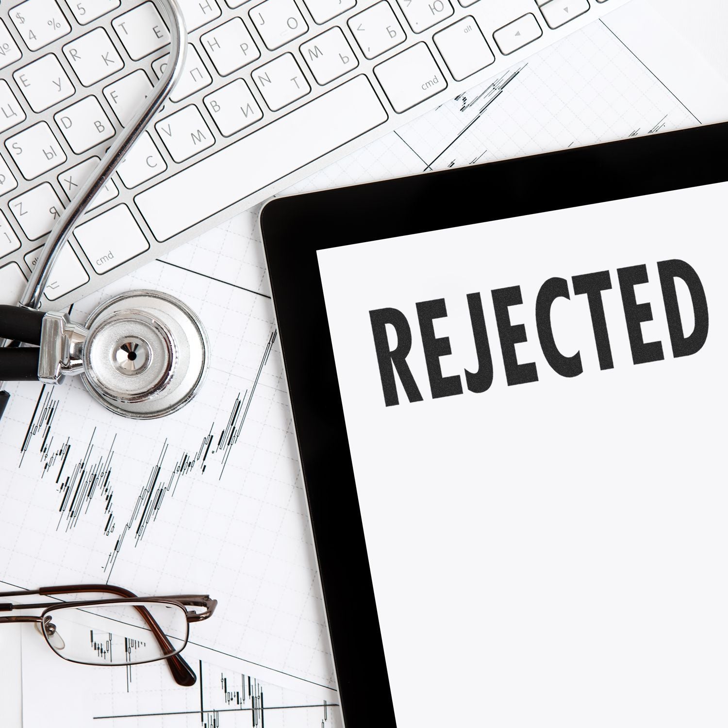 Tablet displaying REJECTED with stethoscope, keyboard, and eyeglasses on a medical chart background, concept of healthcare denial or claim rejection.
