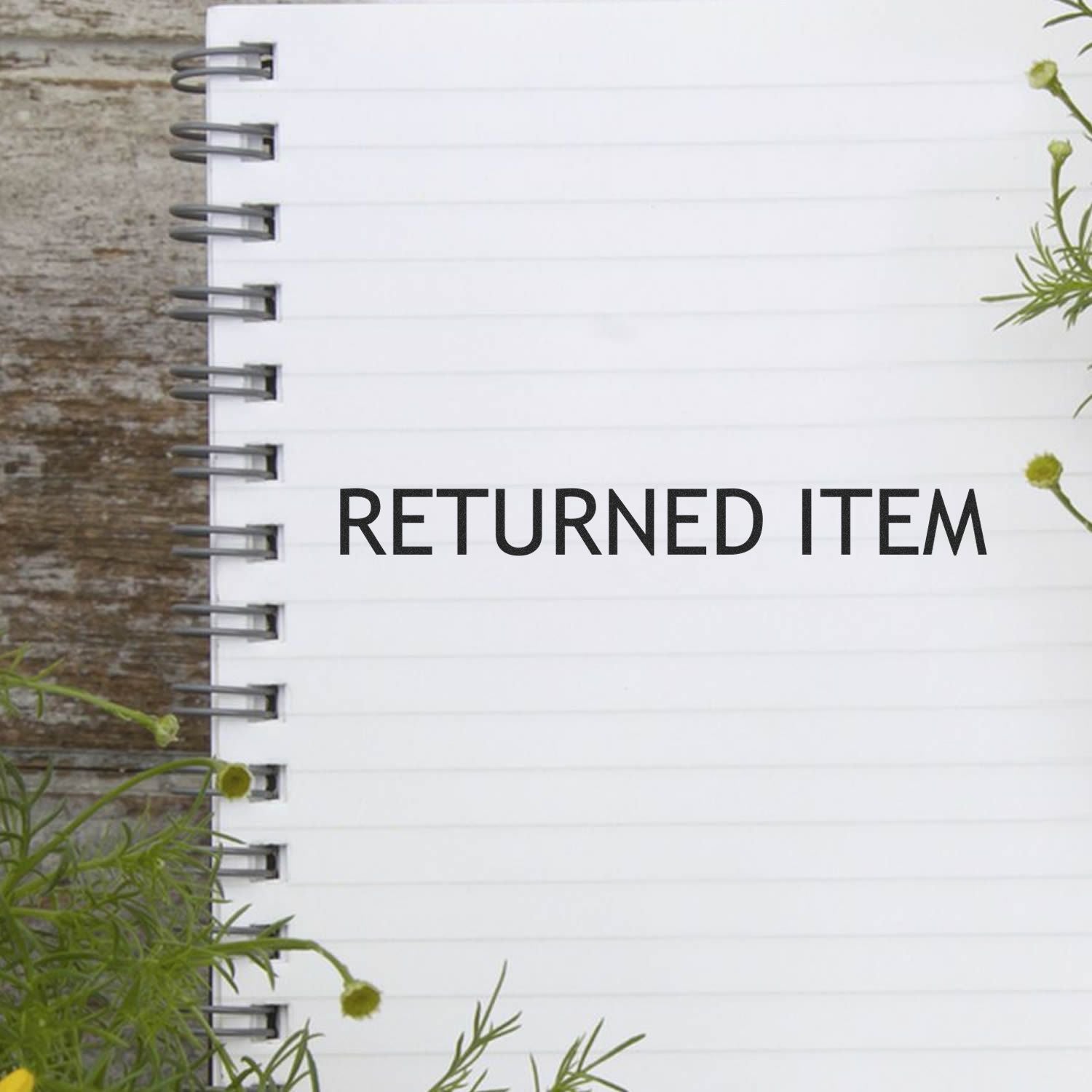 Blank spiral notebook page with RETURNED ITEM text on a rustic wooden table, surrounded by green flowers and foliage.