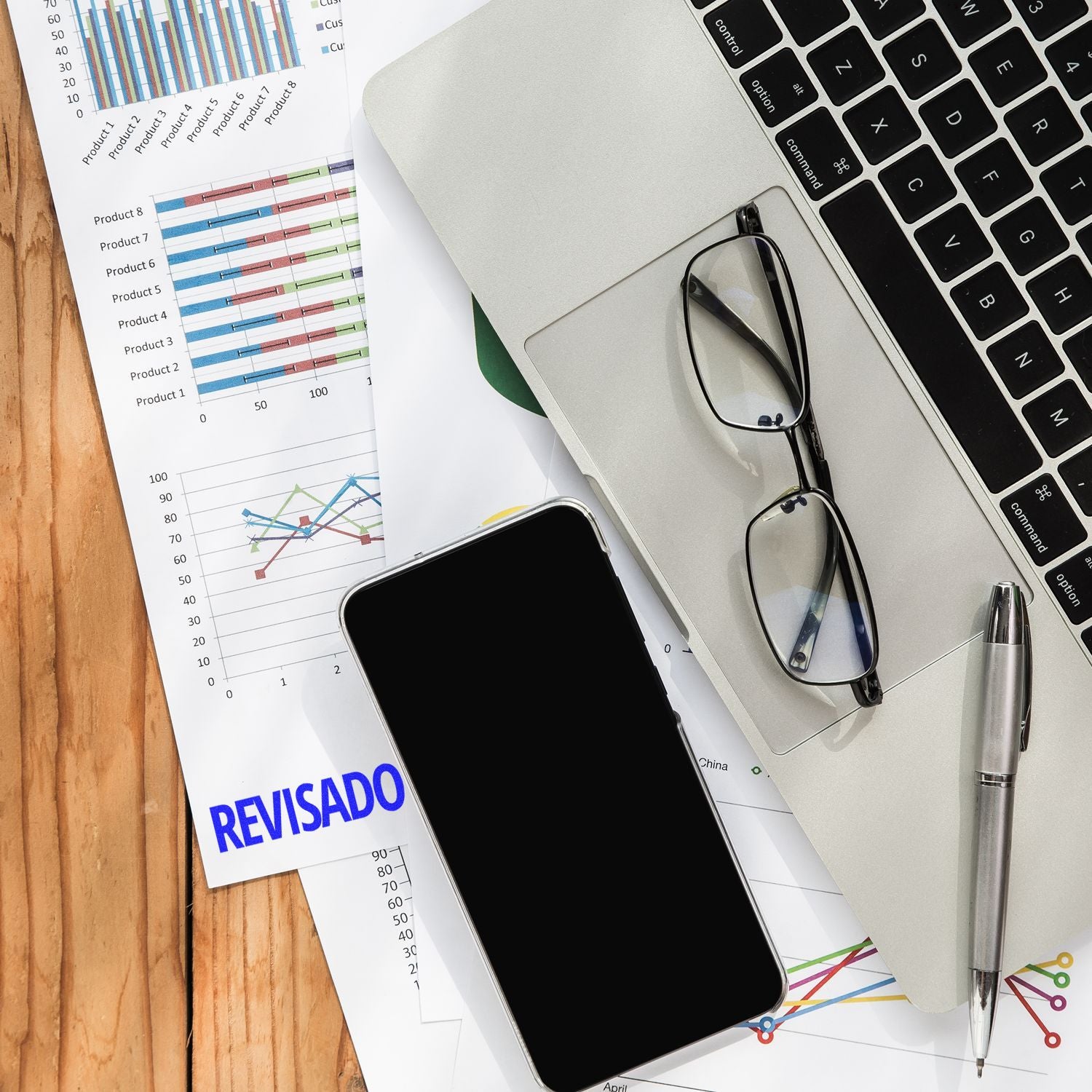 Office desk with laptop, smartphone, eyeglasses, pen, and revised business charts and reports.