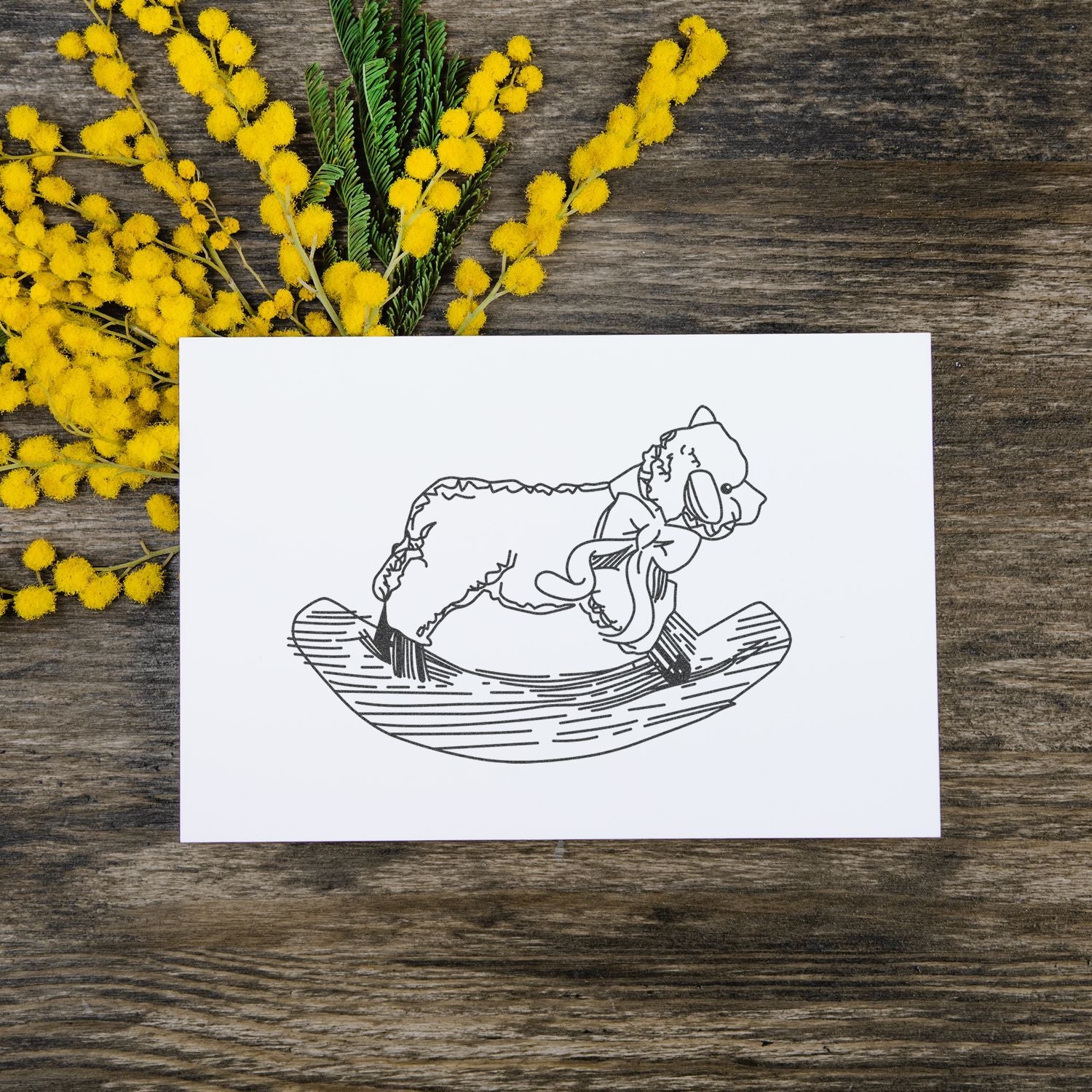 Handmade greeting card with bear illustration on rustic wooden table surrounded by yellow mimosa flowers.