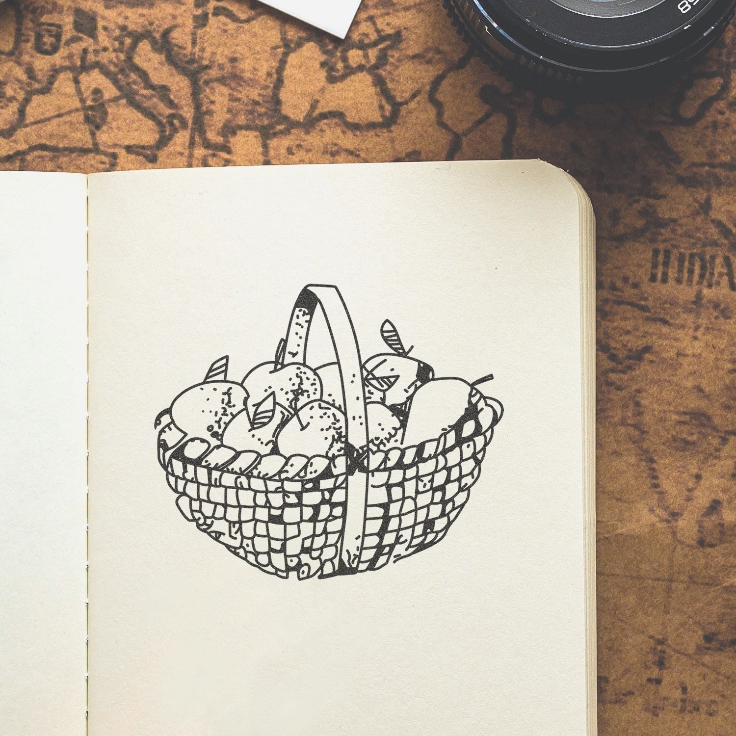 Hand-drawn basket of apples in an open sketchbook on a vintage map background with a camera lens.