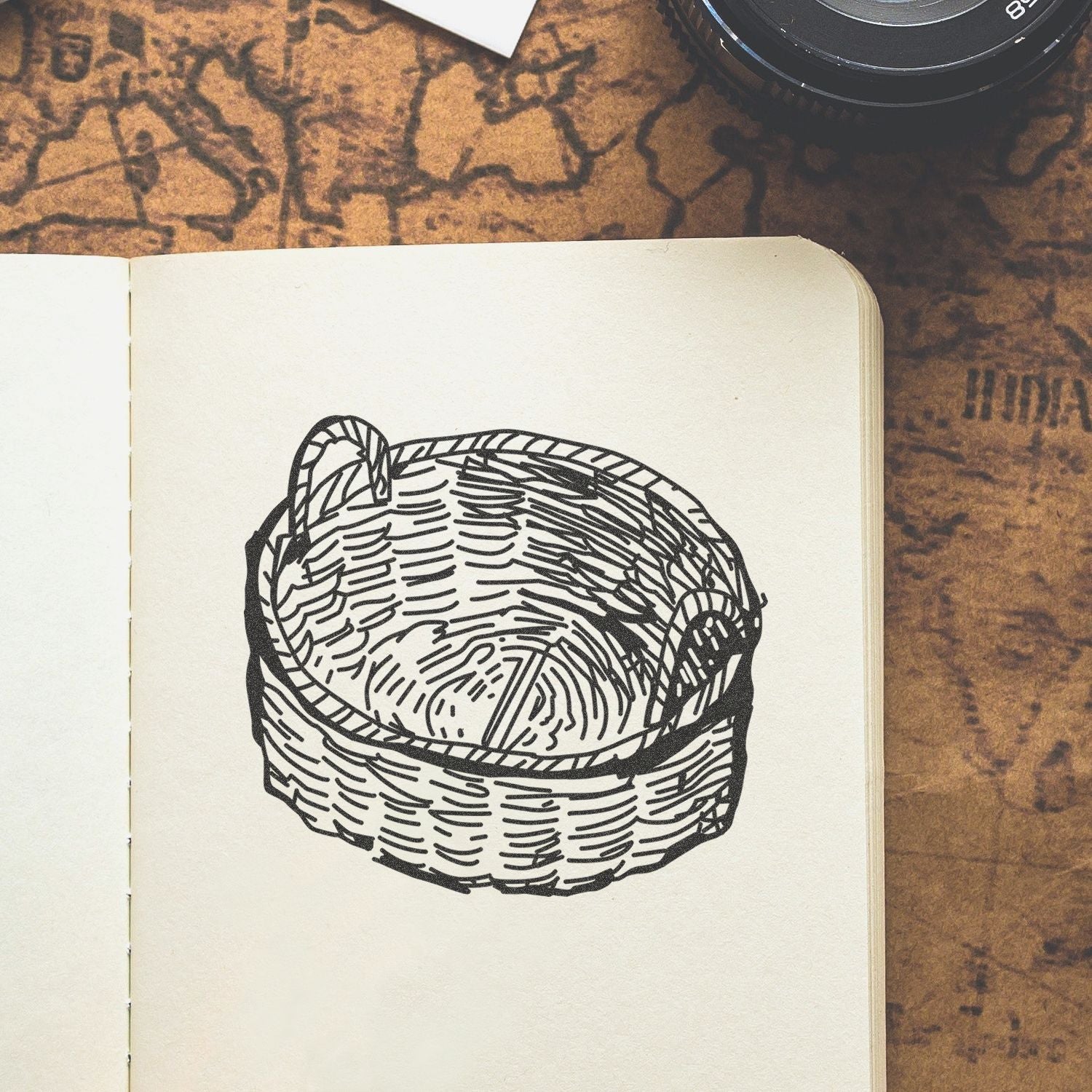 Open sketchbook with black wicker basket illustration on a rustic tabletop, with a camera lens in the corner.