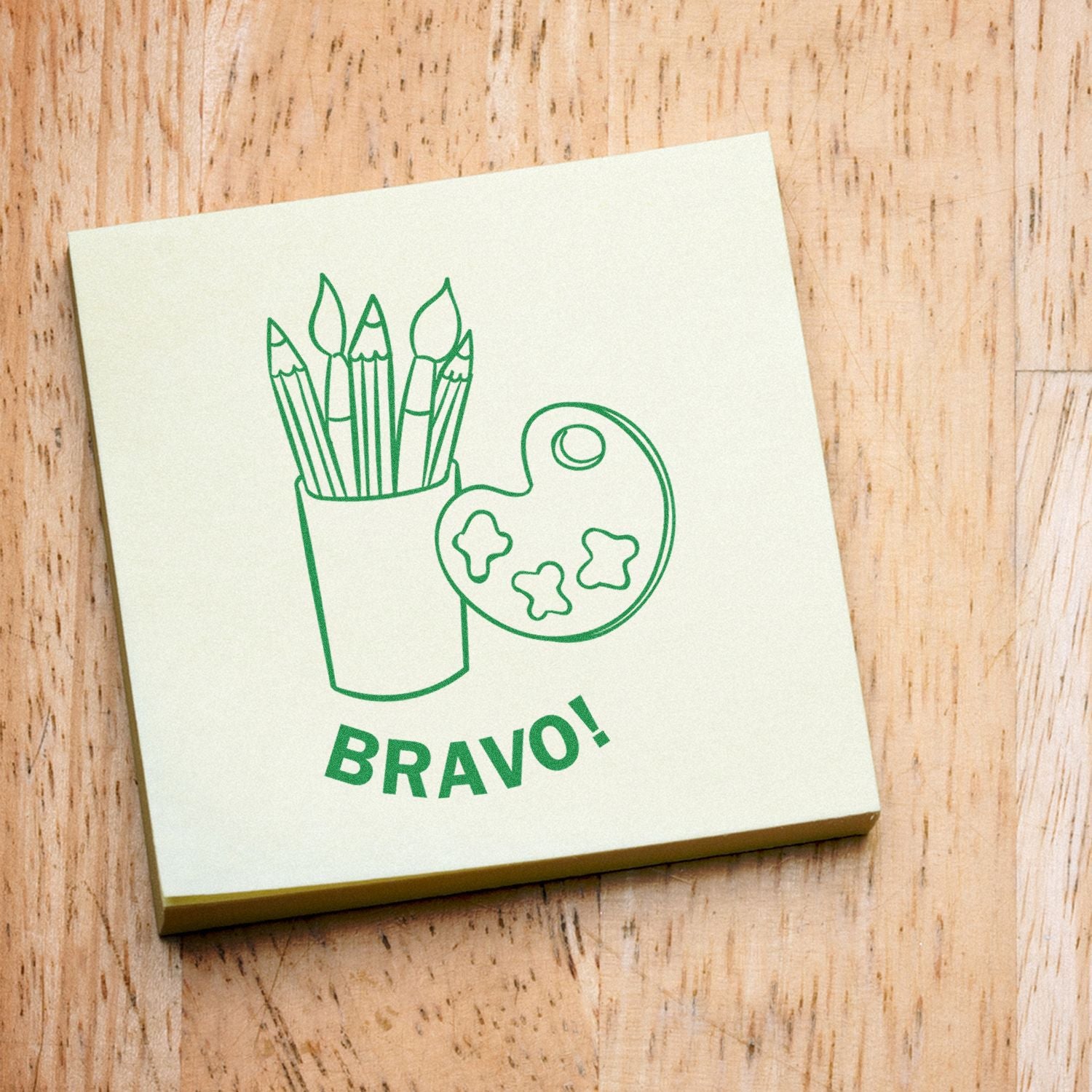 Green BRAVO! sticky note with art supplies illustration, including paintbrushes and palette, on a wooden desk.