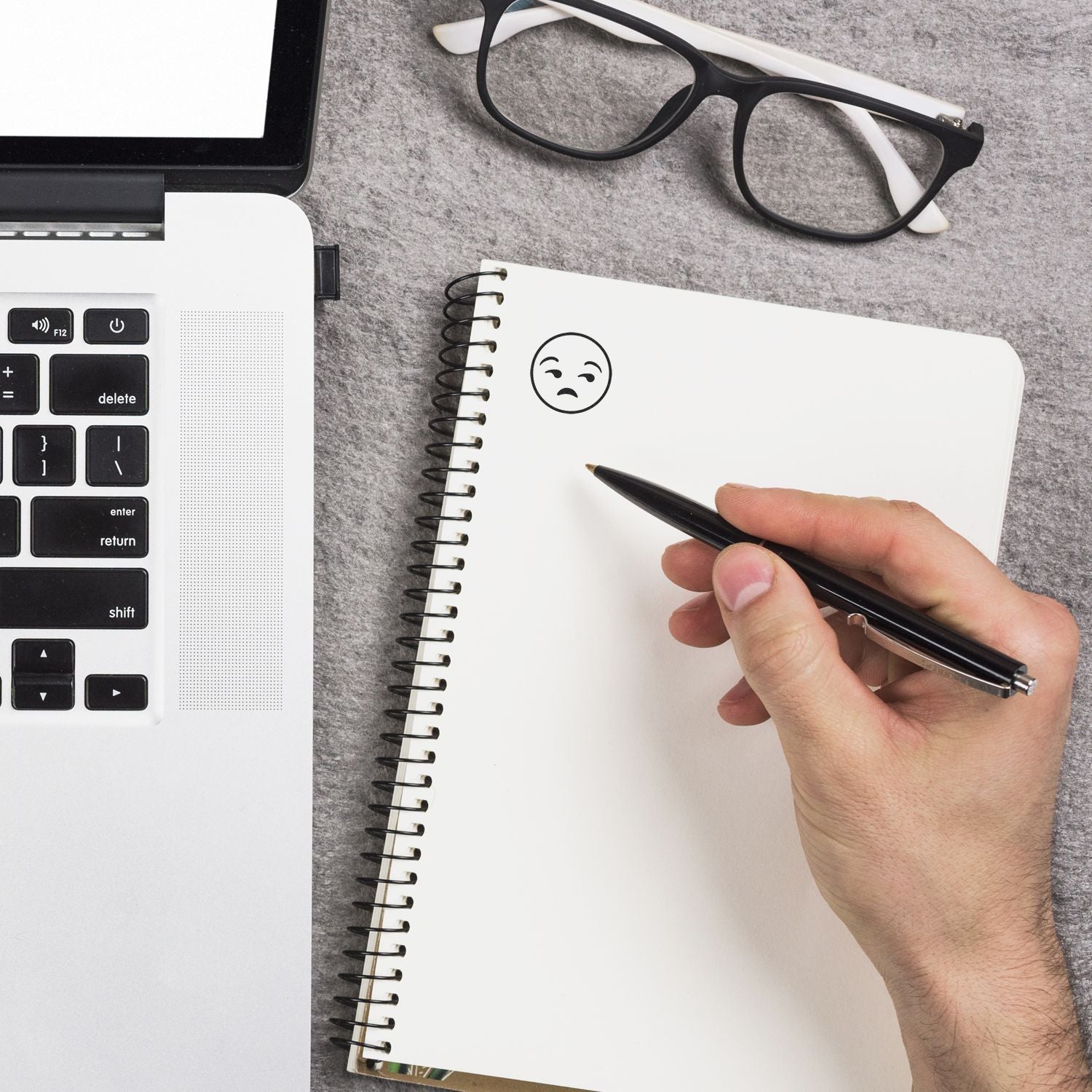 Hand drawing a sad face in a notebook beside a laptop and eyeglasses on a desk, office frustration concept