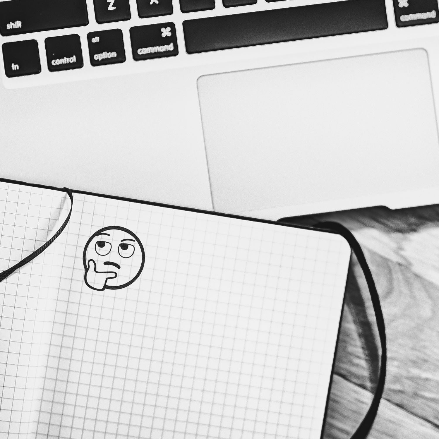 Black and white workspace with laptop keyboard, blank touchpad, and a notebook showing a thinking emoji sketch.